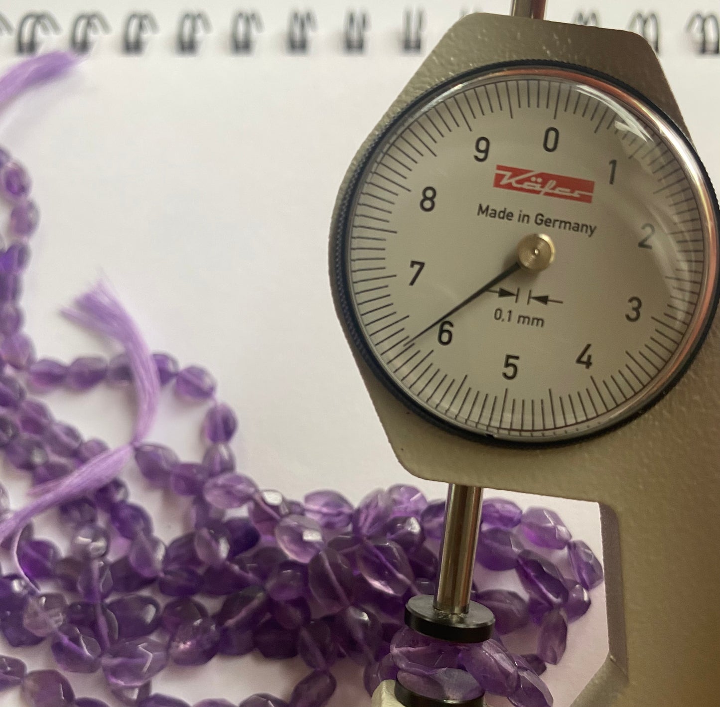 A close-up of a person measuring a purple amethyst faceted oval stone, 5.5mm-to-6.5mm, with dial gauge.