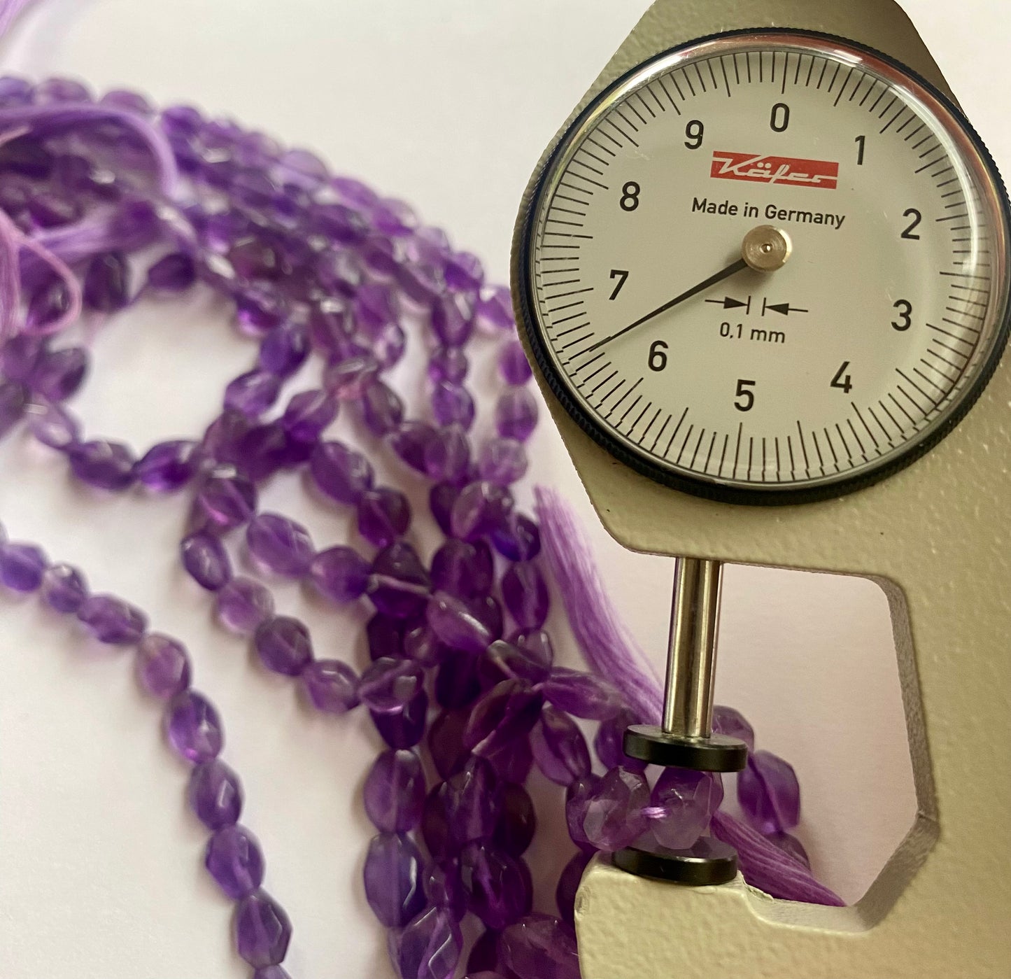 A person measures a purple amethyst faceted oval stone, ranging from 5.5mm-to-6.5mm, using a dial gauge measuring tool.