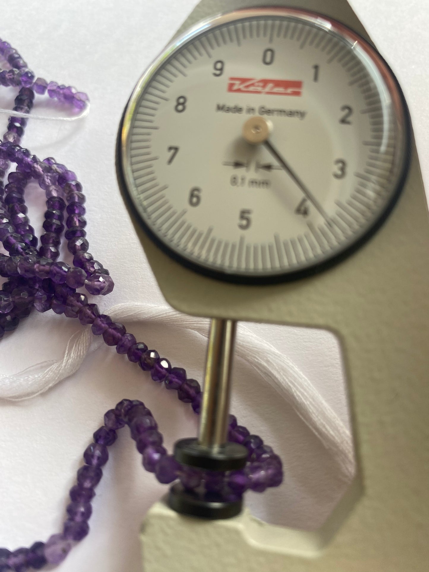 A person measures a purple amethyst faceted rondelle stone, ranging from 4-4.5mm, using a dial gauge measuring tool.