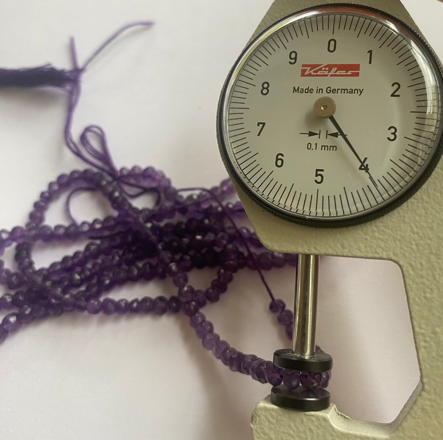 A close-up of a person measuring a purple amethyst faceted rondelle stone, 3.5mm-4mm, with dial gauge.