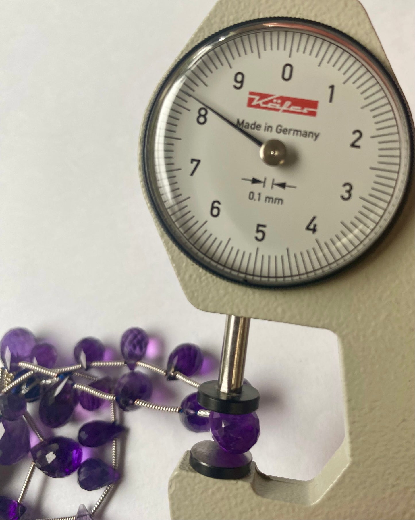 A person measures a purple amethyst teardrop stone, ranging from 5mm to 8mm, using a dial gauge measuring tool on a white background.