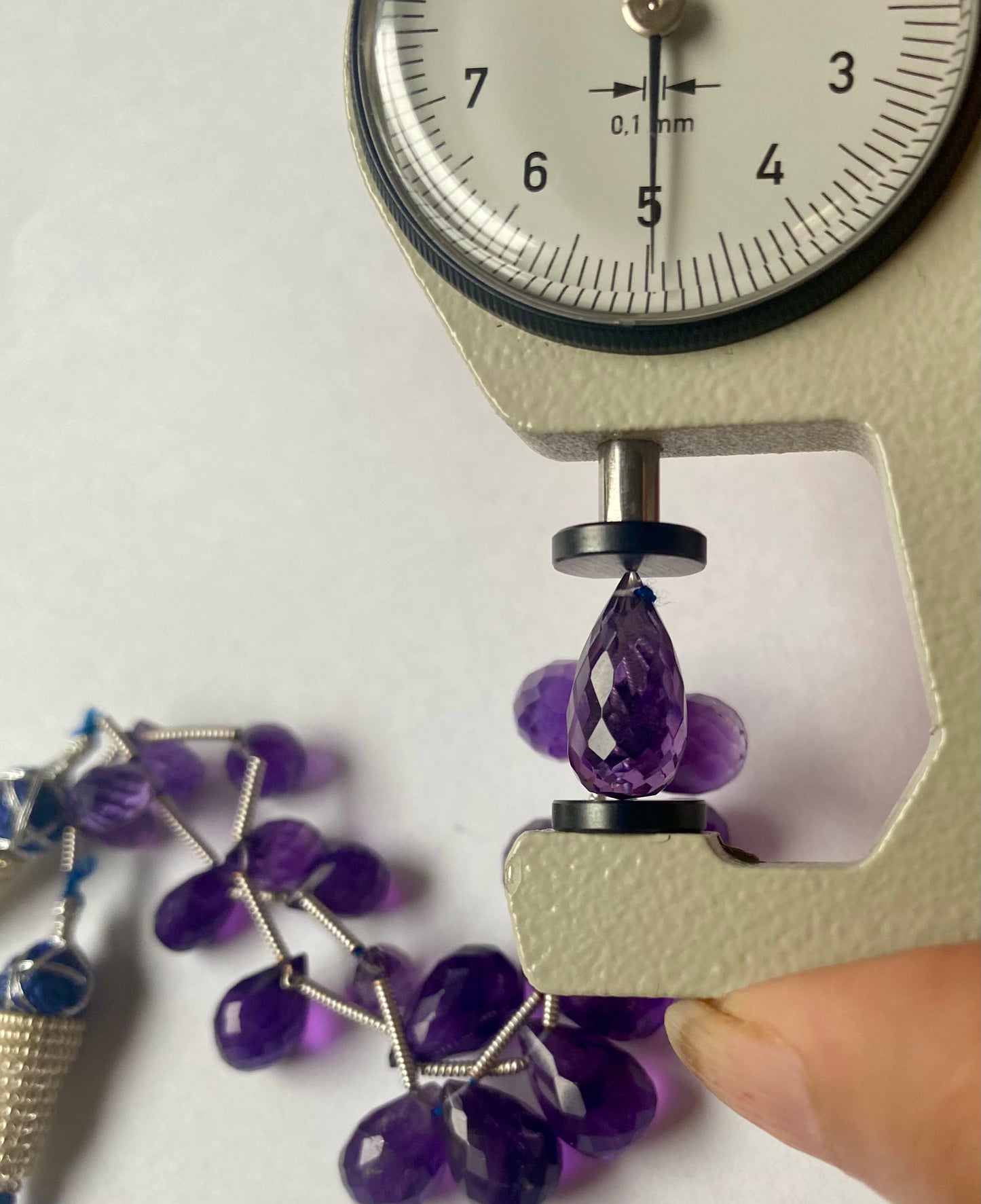 A person measures a purple amethyst teardrop stone, ranging from 5mm to 8mm, using a dial gauge measuring tool.