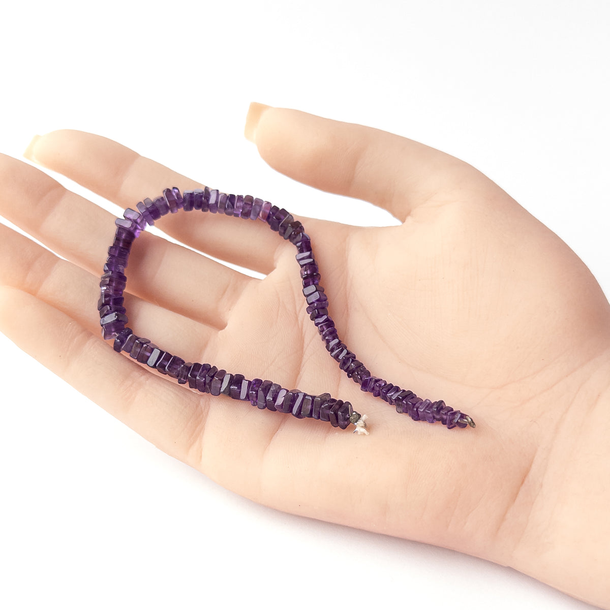 A person holds delicate strands of Amethyst beads, showcasing their smooth square heishi design.
