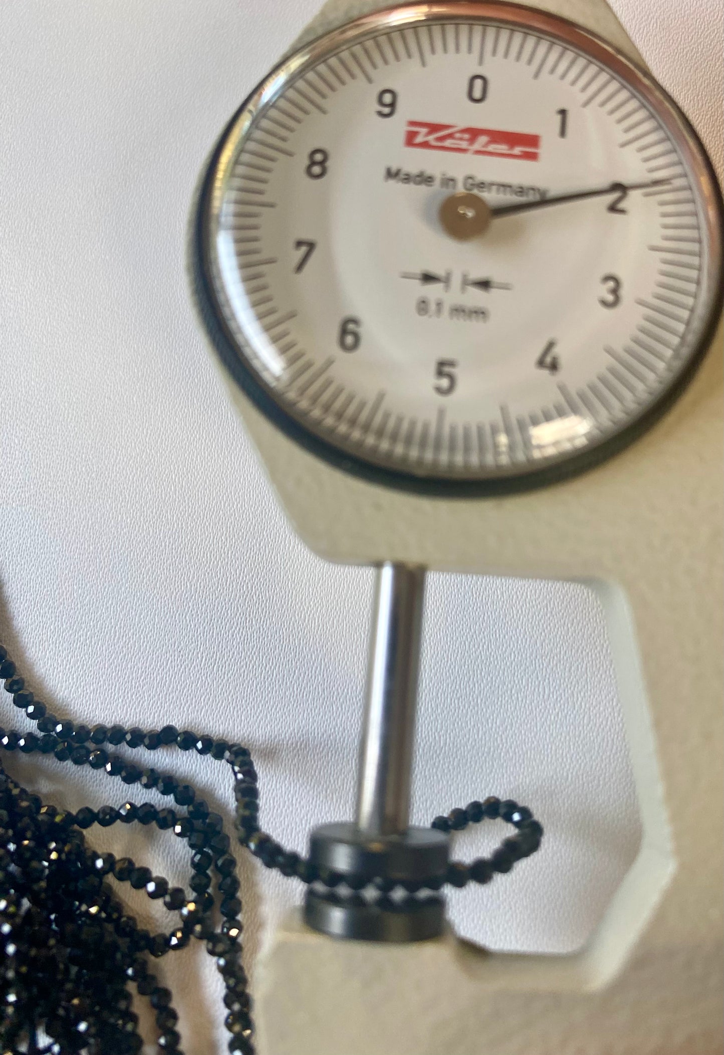 A dial gauge displaying a black bead beside a black gemstone strand made of micro-faceted rondelle spinel beads, 2-2.5mm in size.