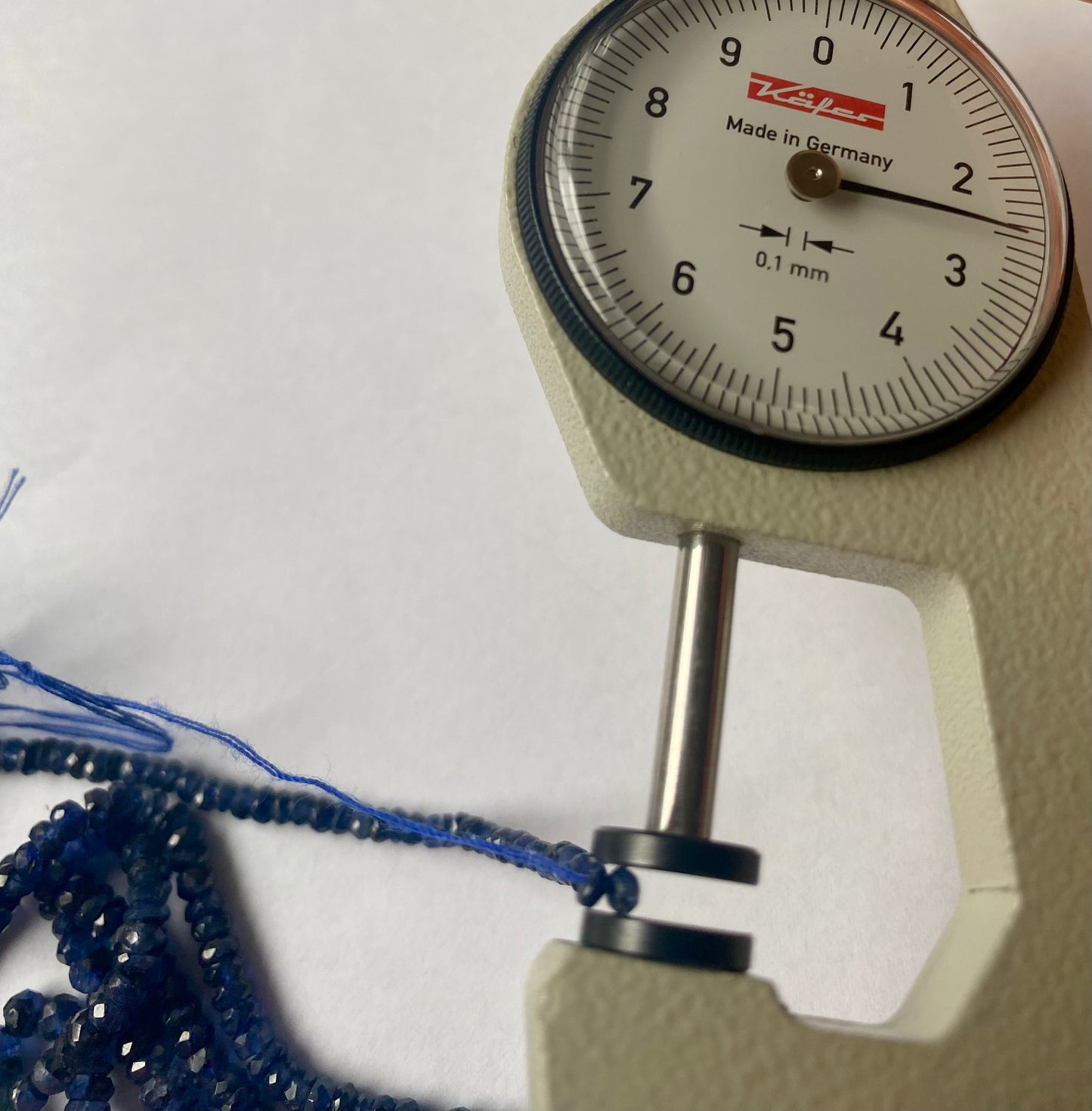 Close-up of a person using a measuring dial gauge to measure a blue sapphire faceted rondelle bead, highlighting its dimensions.