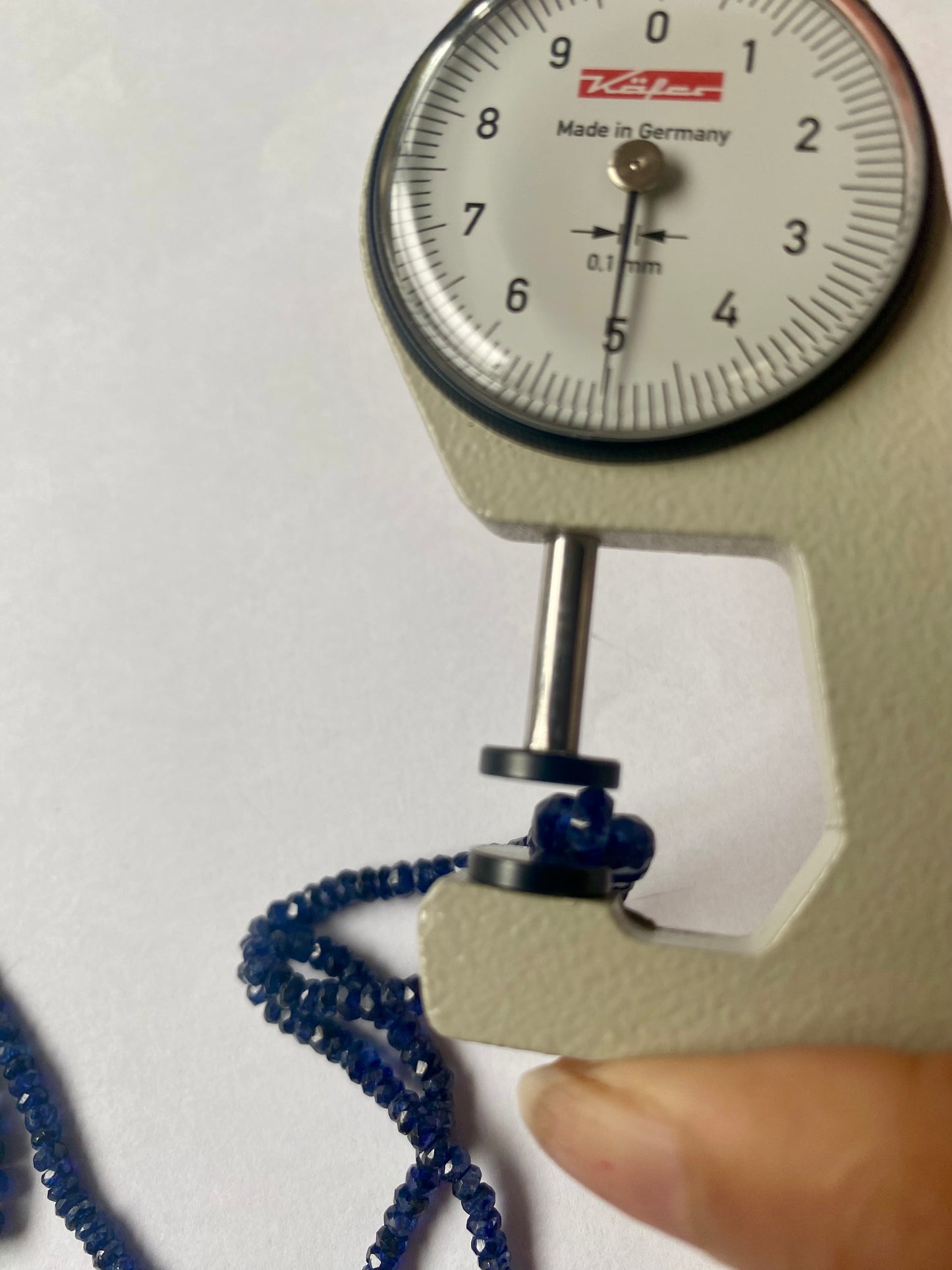 A person measuring a blue sapphire faceted rondelle bead with a dial gauge measure, showcasing its size and detail.