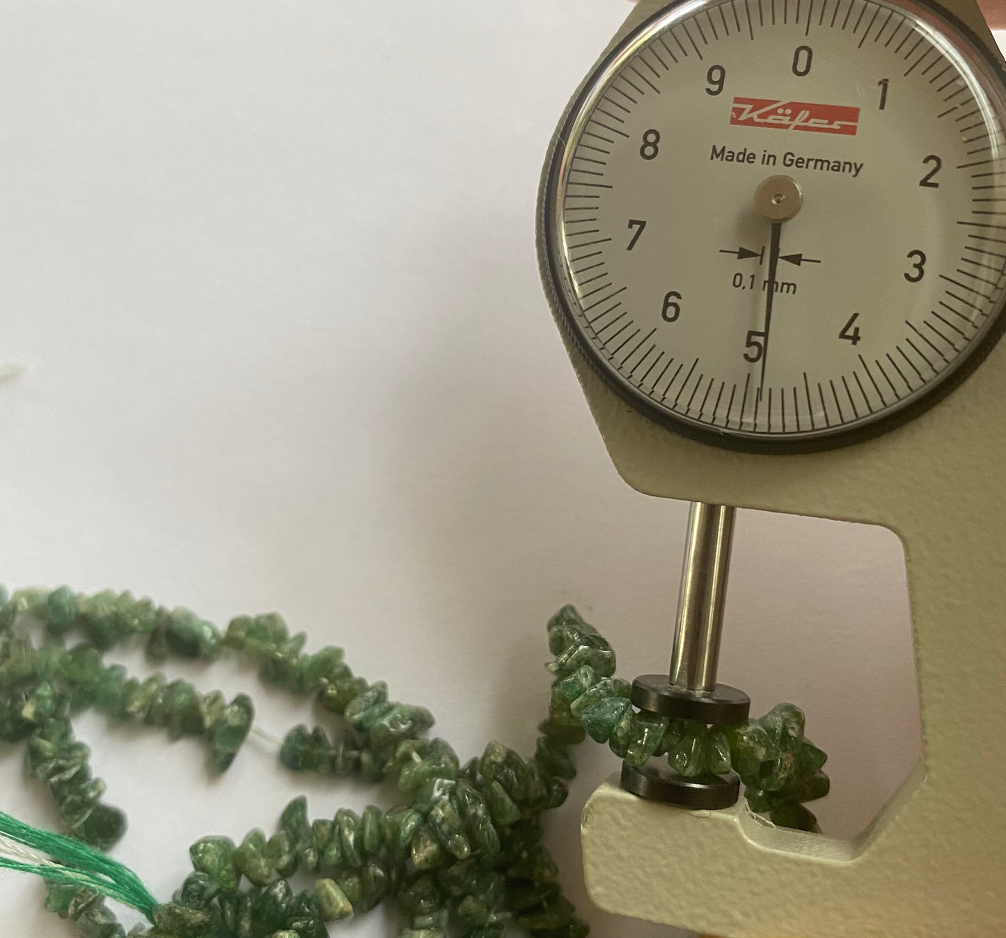 A person measures a green emeral chip stone, ranging from 5mm to 7mm, using a dial gauge measuring tool.