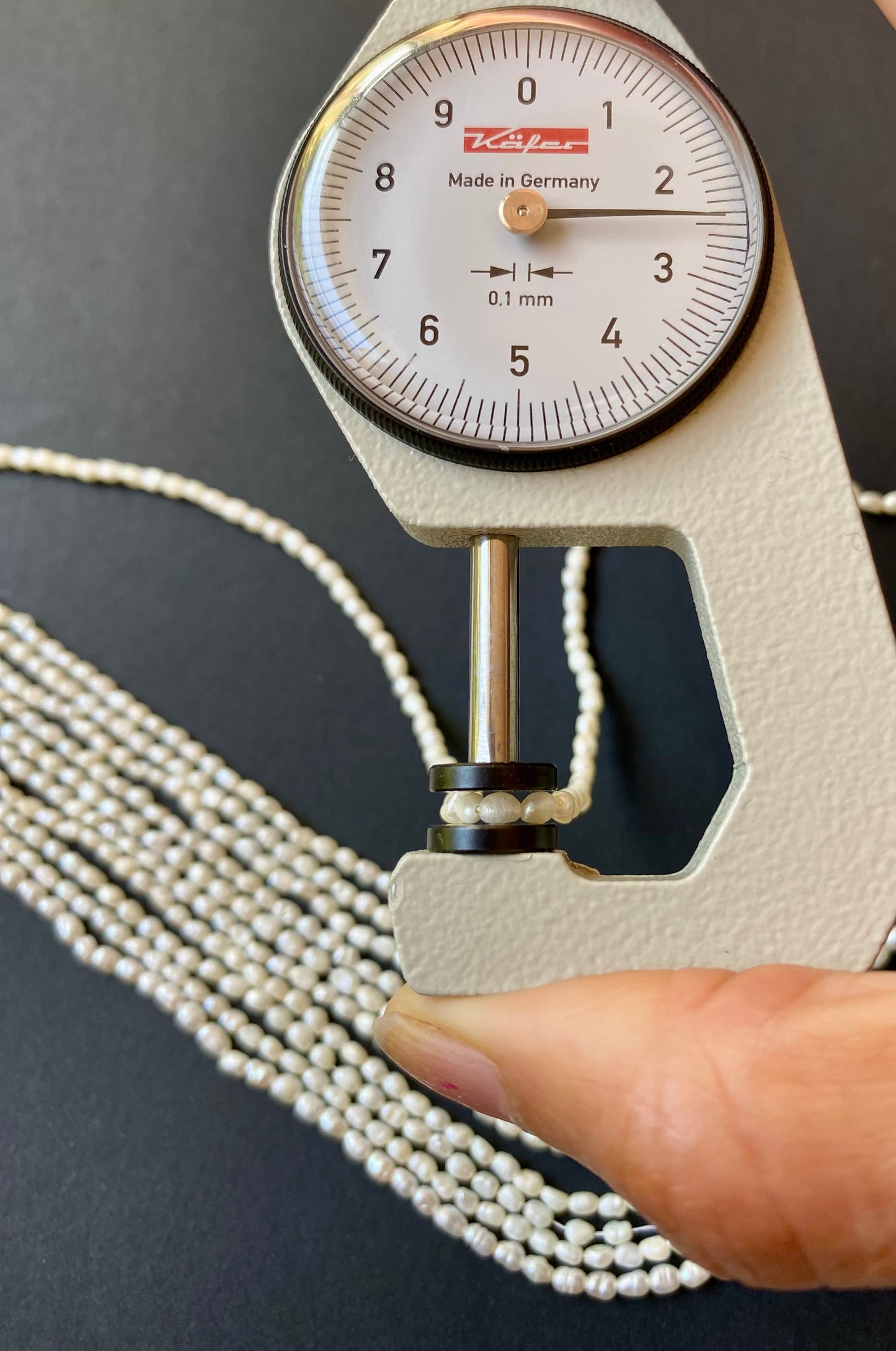 A hand holds a measuring dial gauge next to a strand of freshwater rice pearls, showcasing their 2-2.5mm bead size.