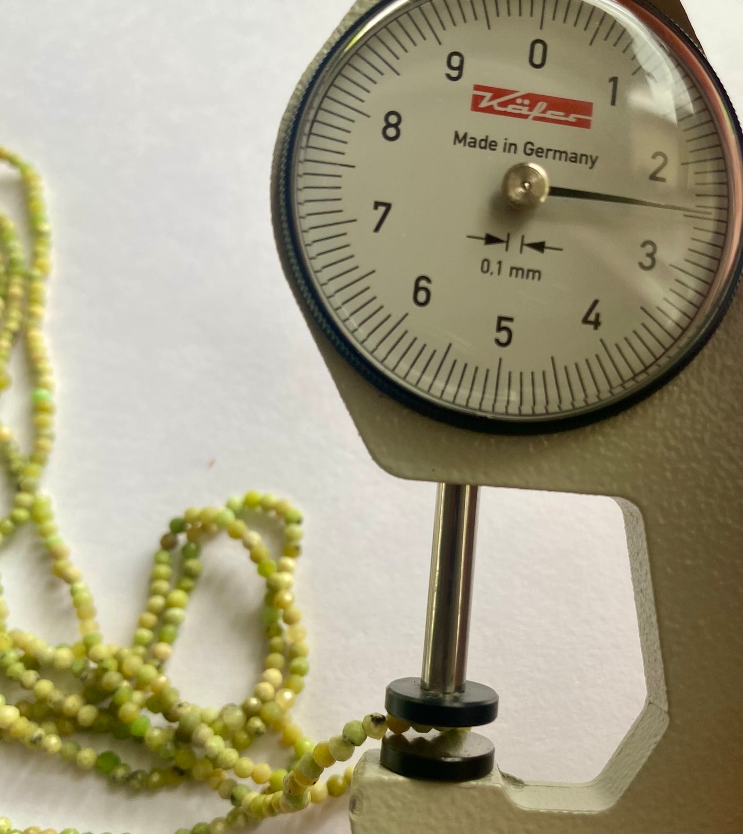 A dial gauge with a green bead on it next to a green gemstone strand featuring 2.5mmfaceted rondelle green opal beads.