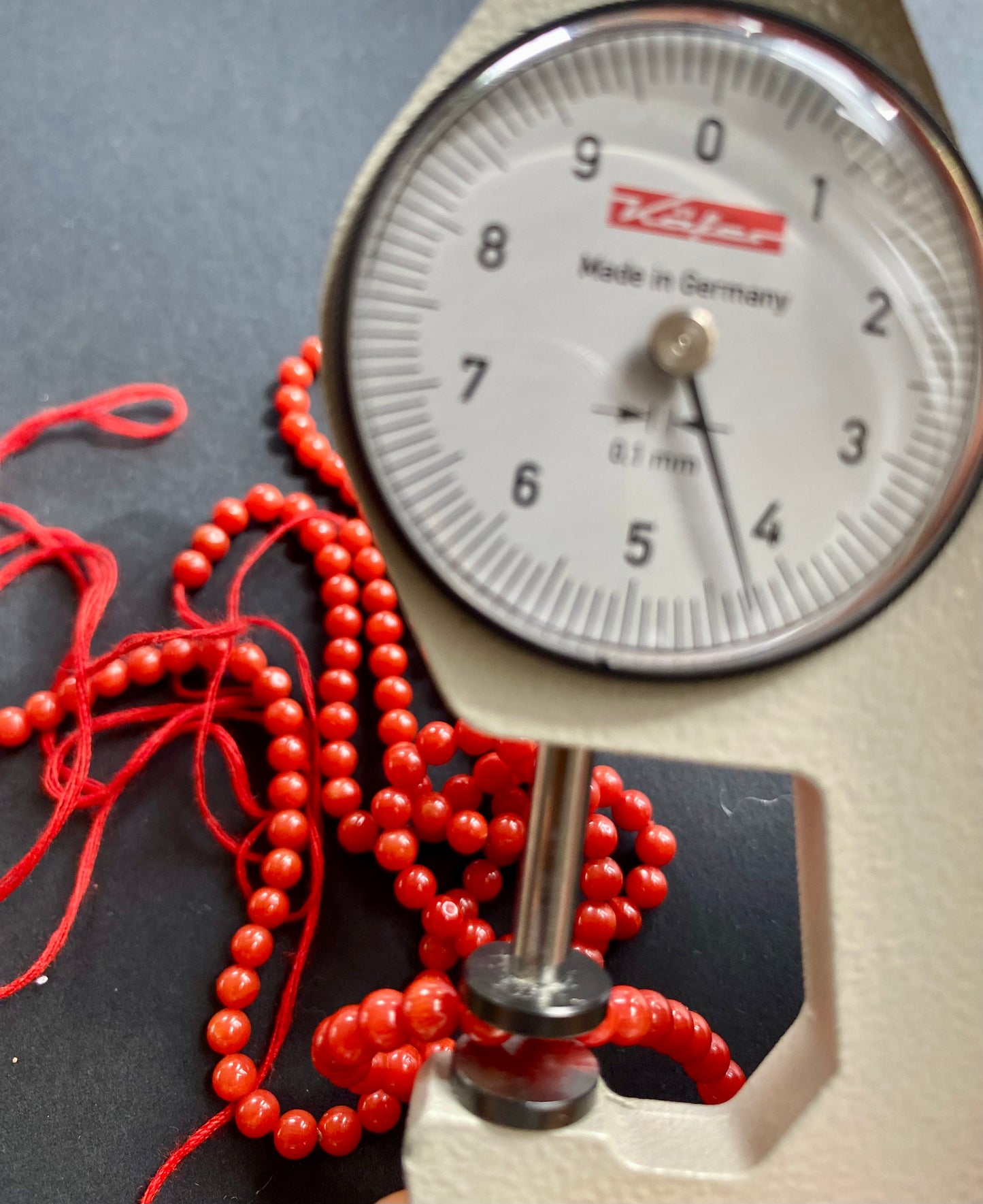 Close-up of a measuring dial gauge alongside smooth, round 4mm Italian coral beads in vibrant red.