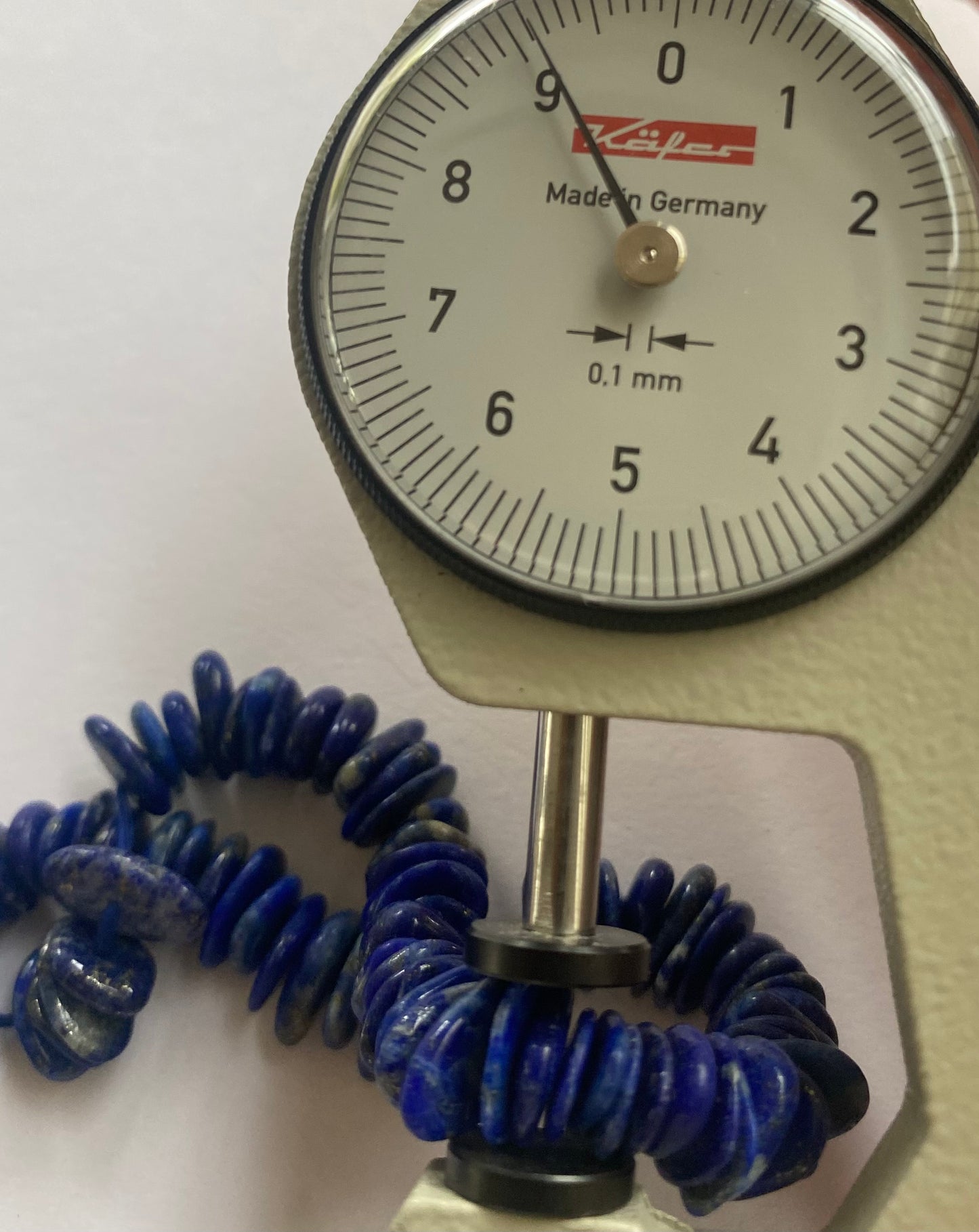 Close-up of a measuring dial gauge tool alongside a string, highlighting the precision of measurements for Lapis Heishi Smooth beads.