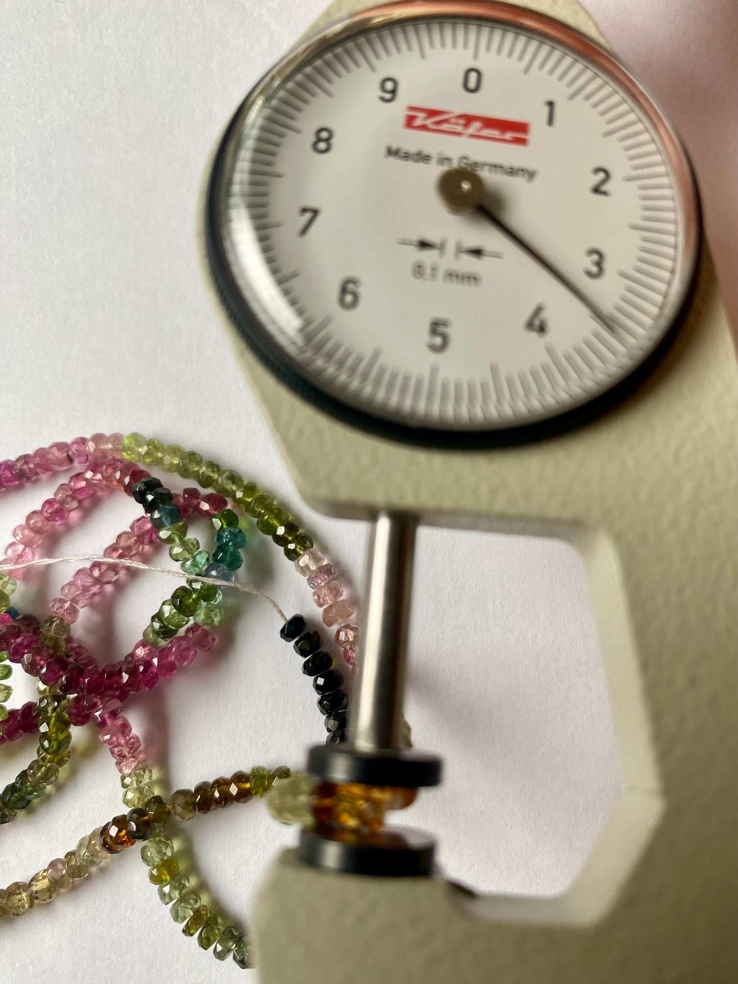 Close-up of a measuring dial gauge alongside a vibrant collection of 3.5mm micro-cut tourmaline beads.