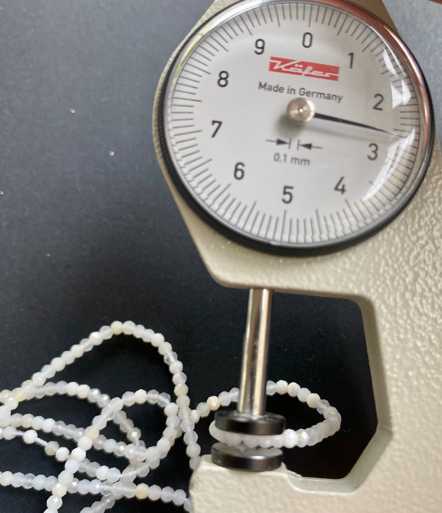 A dial gauge displaying a bead beside a black gemstone strand made of micro-faceted rondelle opal beads, 2.6-2.7mm in size.