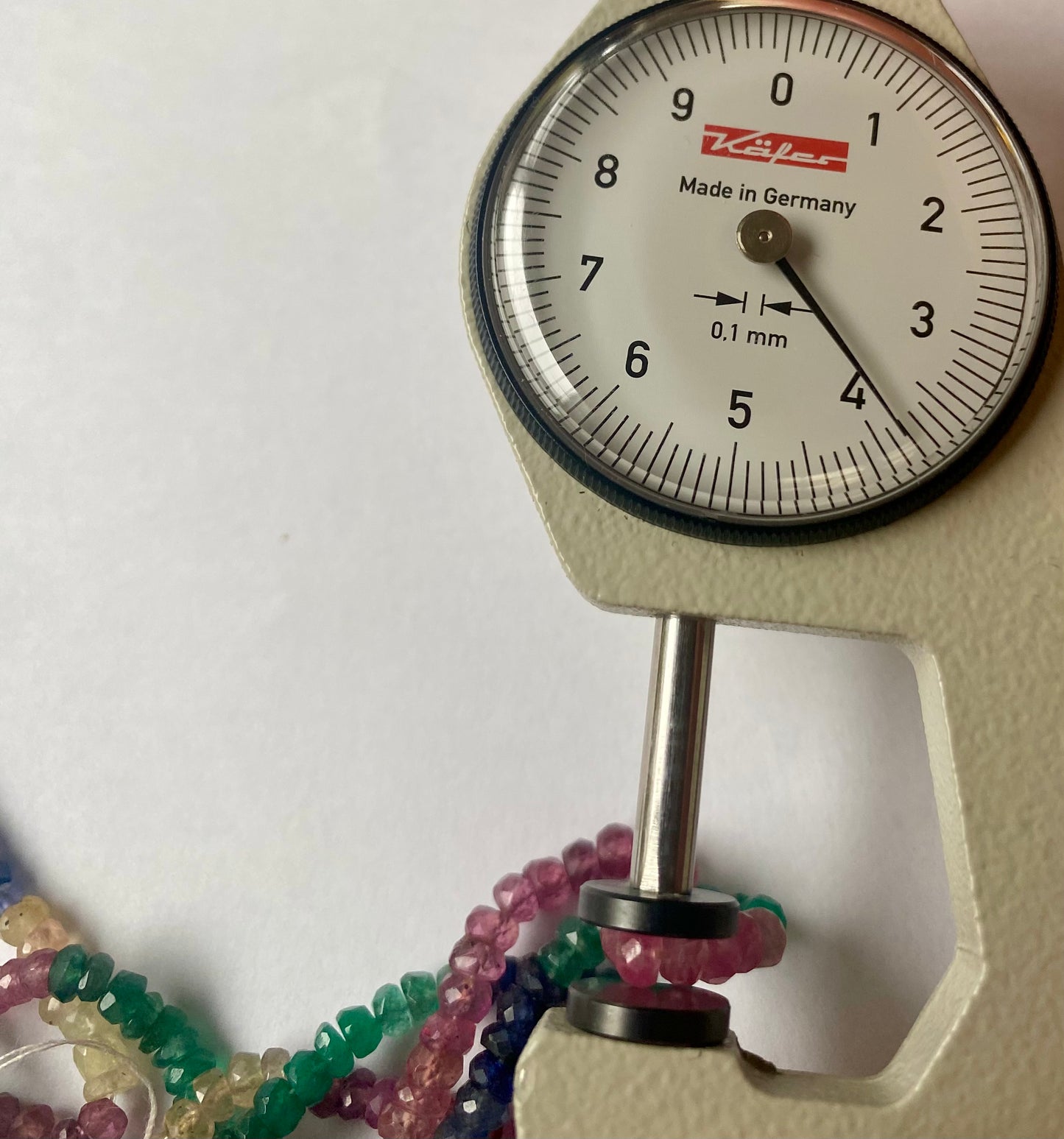 Close-up of a measuring dial gauge beside colourful multi-precious beads, including ruby, blue sapphire, yellow sapphire, and emerald.