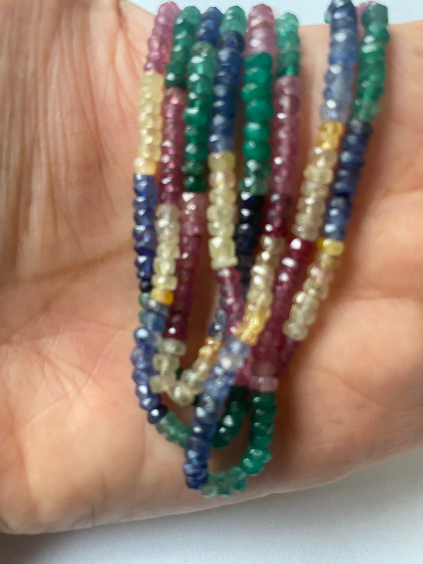 A person holds a vibrant bunch of multi-coloured faceted beads, including ruby, blue sapphire, yellow sapphire, and emerald.