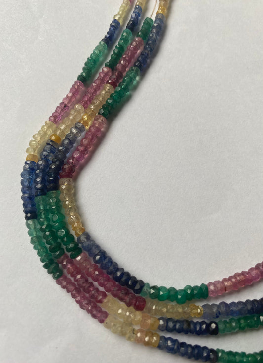 A vibrant multi-coloured necklace featuring ruby, blue sapphire, yellow sapphire, and emerald beads on a white background.