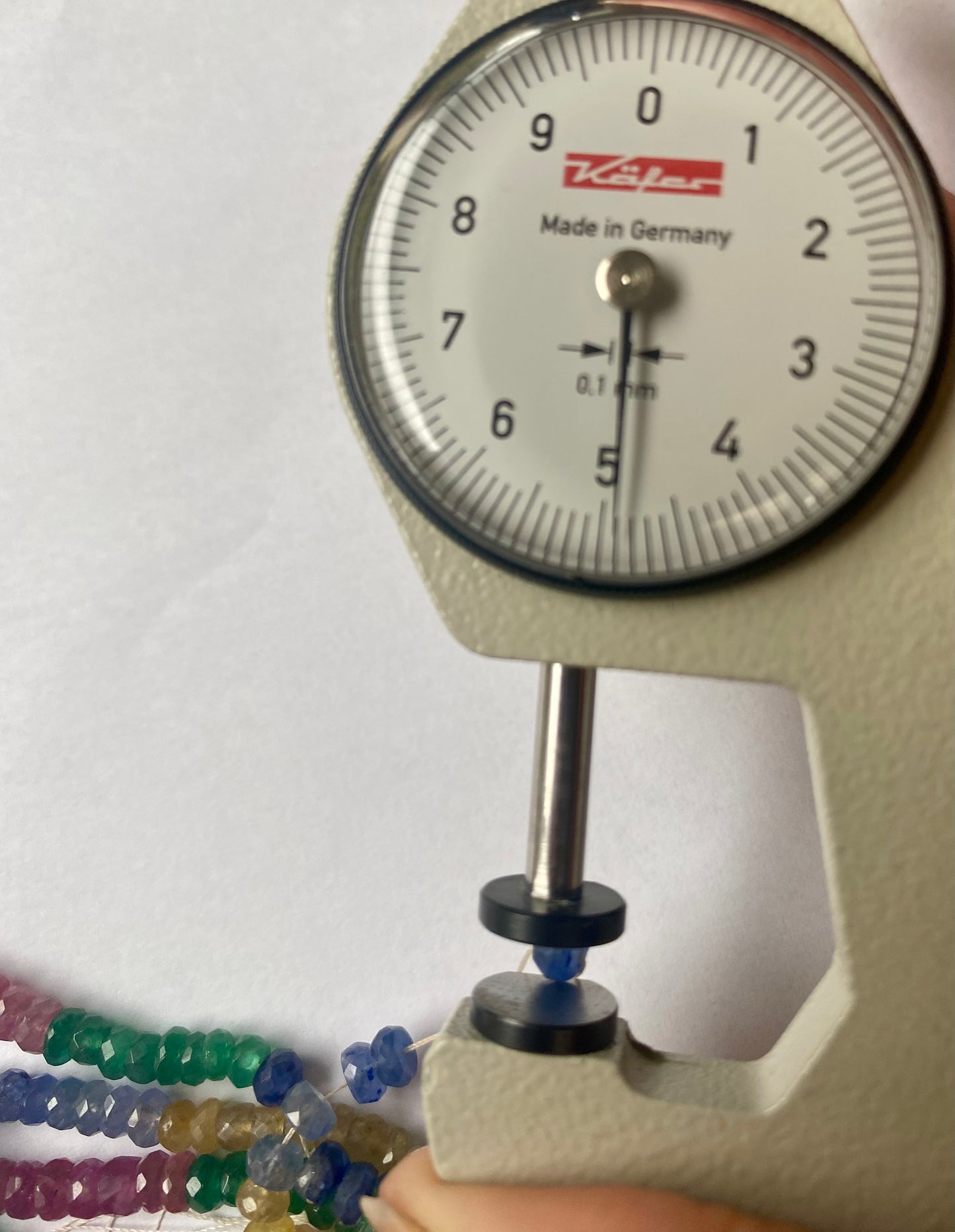 Close-up of a measuring dial gauge beside colourful multi-precious beads, including ruby, blue sapphire, yellow sapphire, and emerald.