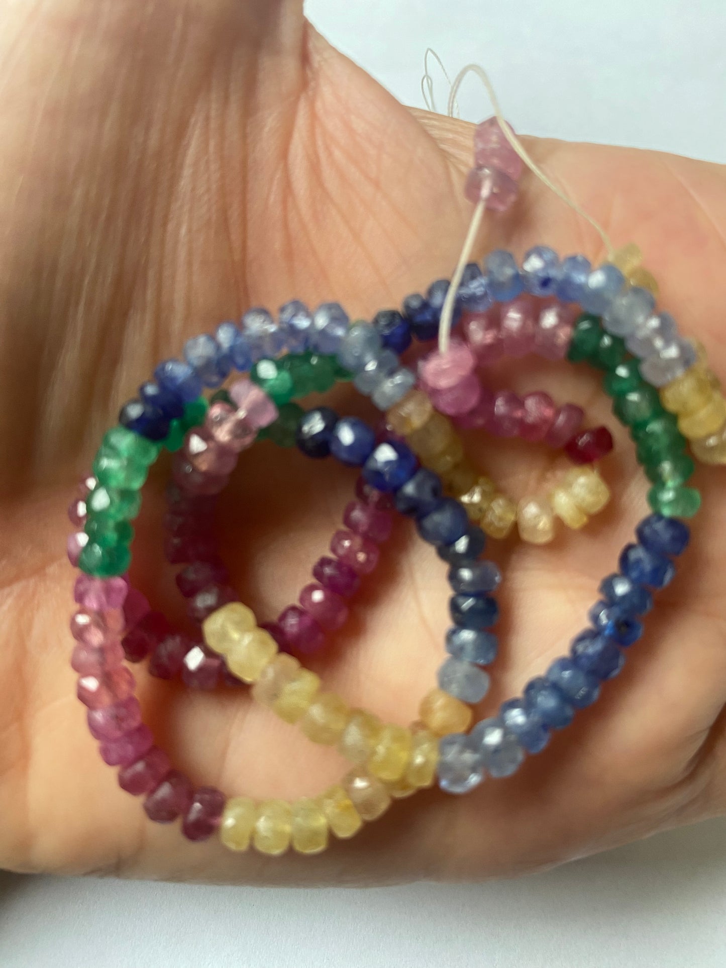 A person holds a vibrant bunch of multi-coloured faceted beads, including ruby, blue sapphire, yellow sapphire, and emerald.