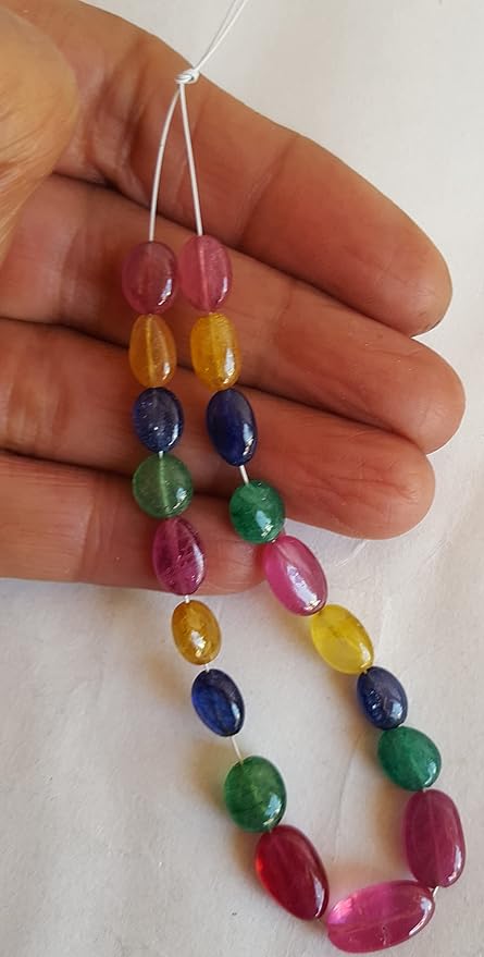 A person holds a vibrant bunch of multi-coloured smooth oval beads, including ruby, blue sapphire, yellow sapphire, and emerald.