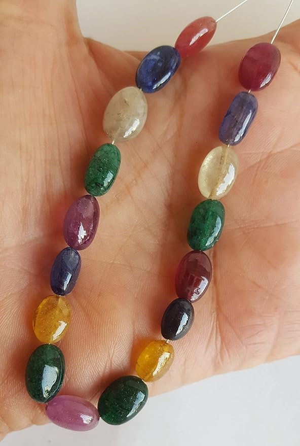 A person holds a vibrant bunch of multi-coloured smooth oval beads, including ruby, blue sapphire, yellow sapphire, and emerald.