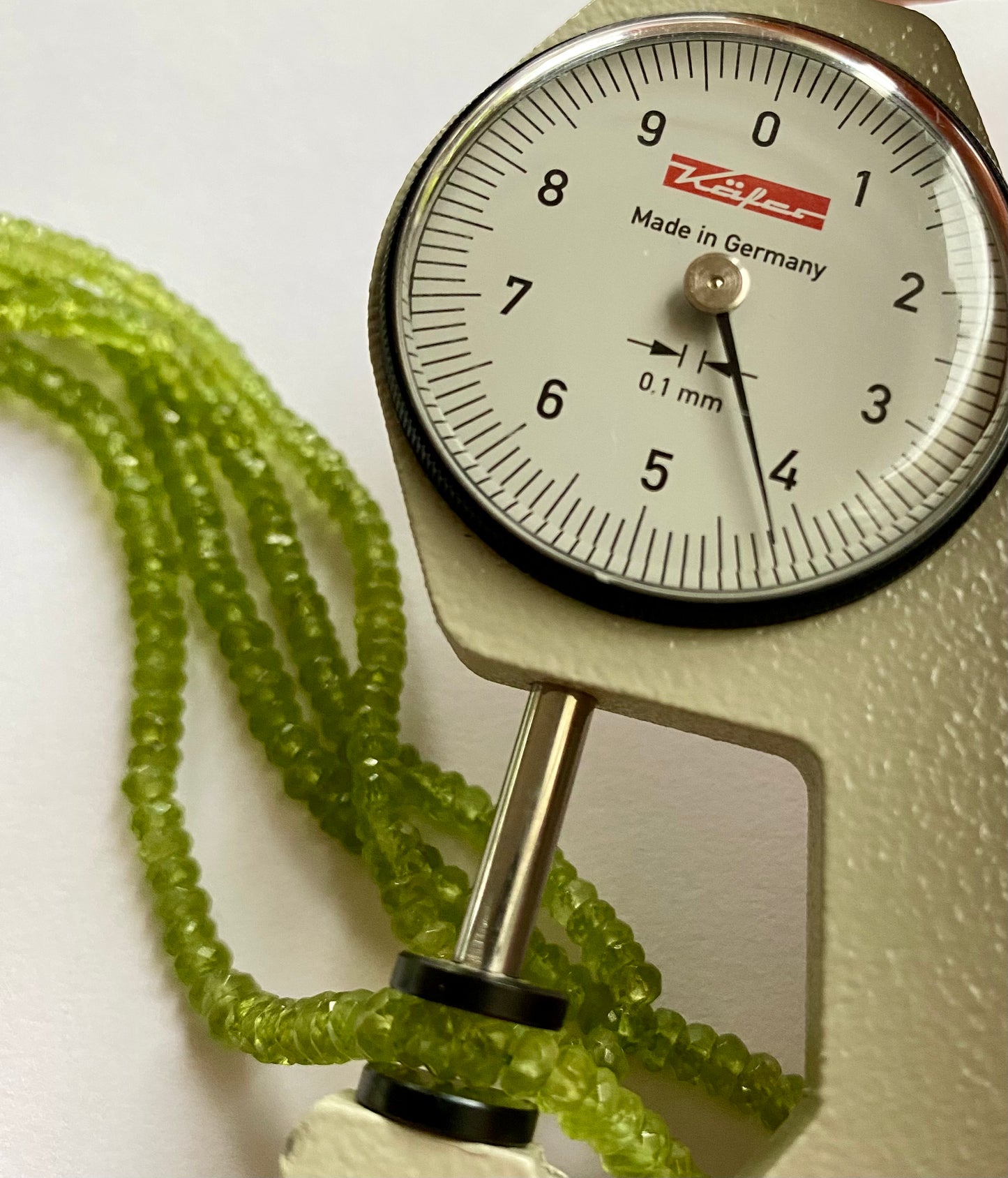 A person measuring a peridot faceted rondelle bead with a dial gauge measure, showcasing its size and detail.