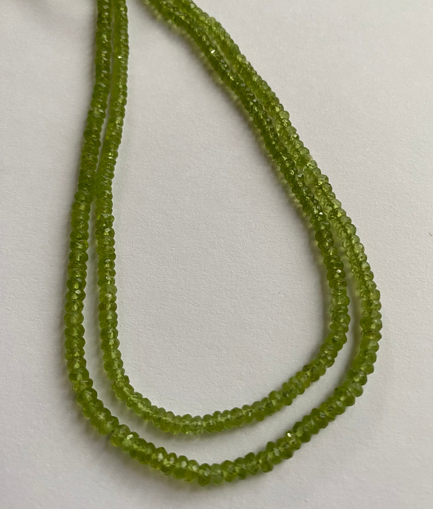 A close-up of a multiple strands of green peridot gemstone beads, faceted rondelles ranging from 3.7mm to 4.4mm, glistening in the light.