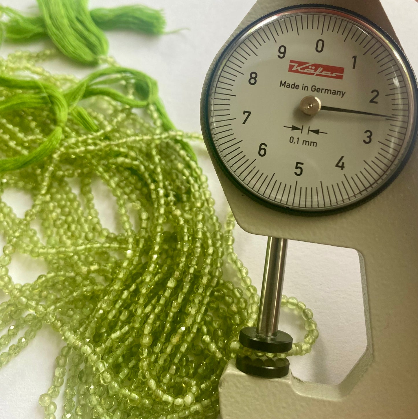 A person measures a green peridot smooth rondelle stone, ranging from 2.5mm to 2.8mm, using a dial gauge measuring tool.