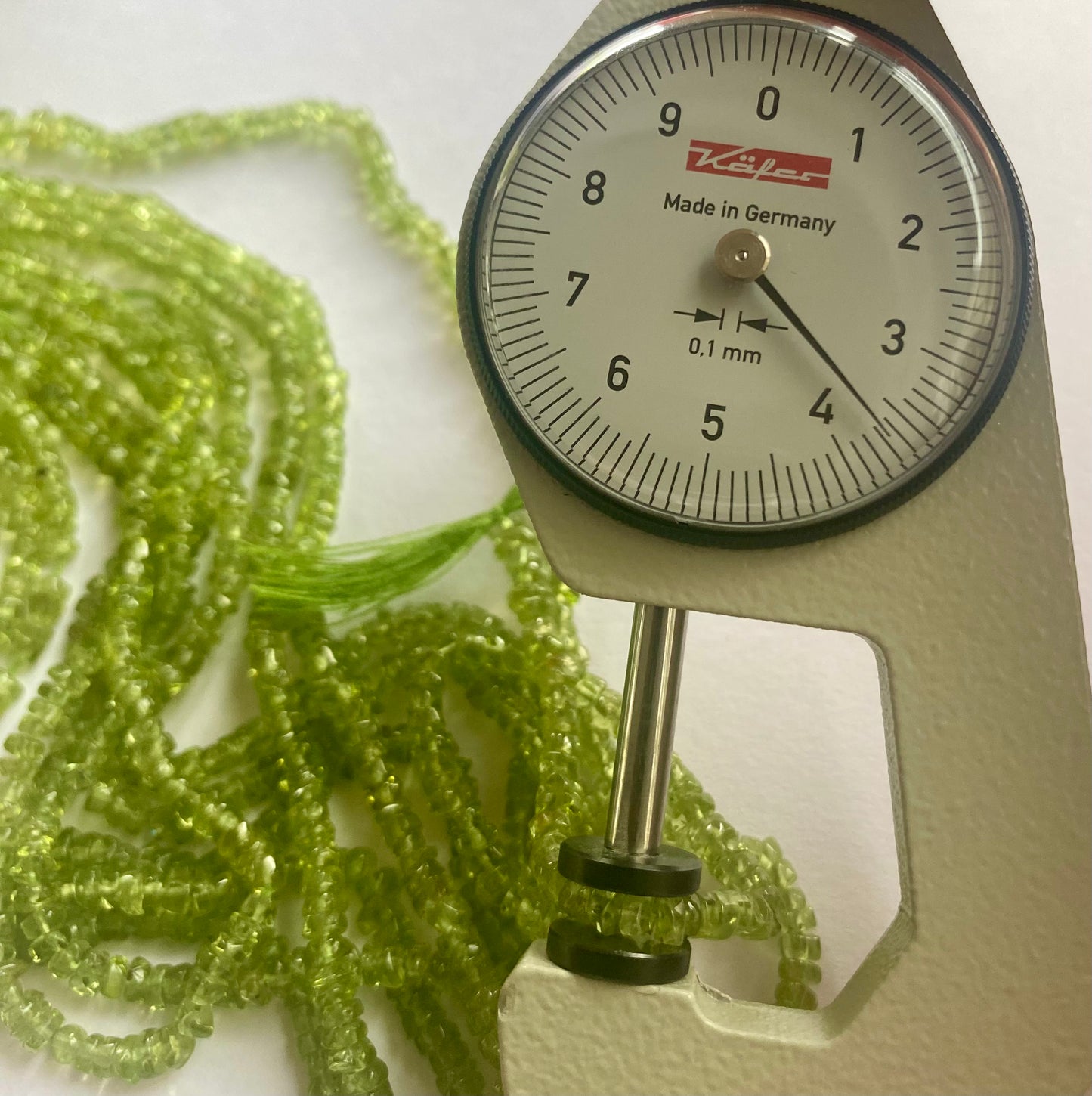 Close-up of a measuring dial gauge alongside smooth green peridot beads, ranging from 3.5mm to 4mm in size.