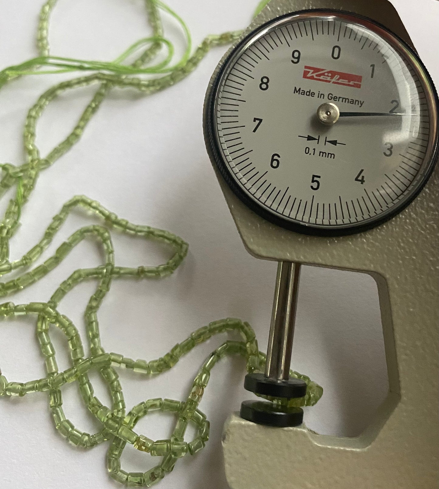 A person measures a green peridot tube shaped stone, ranging from 2mm to 2.2mm, using a dial gauge measuring tool.