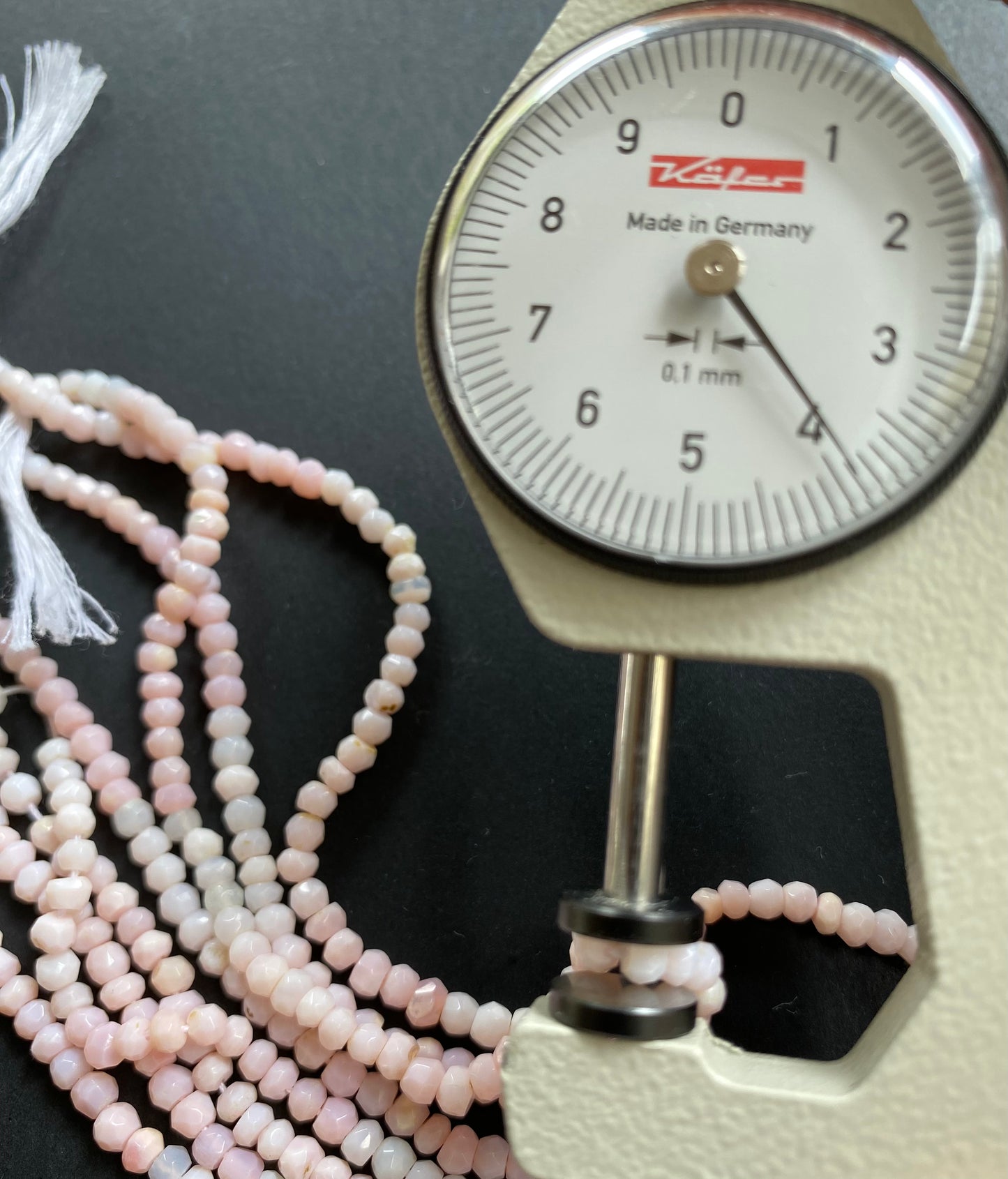 Close-up of a person using a measuring dial gauge to measure a Pink Opal faceted rondelle bead, highlighting its dimensions.