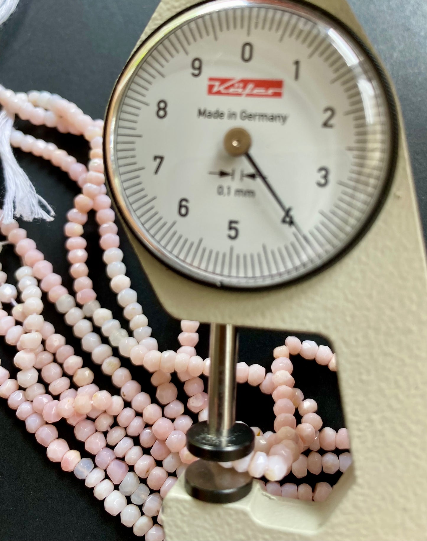 A person measuring a Pink Opal faceted rondelle bead with a dial gauge measure, showcasing its size and detail.