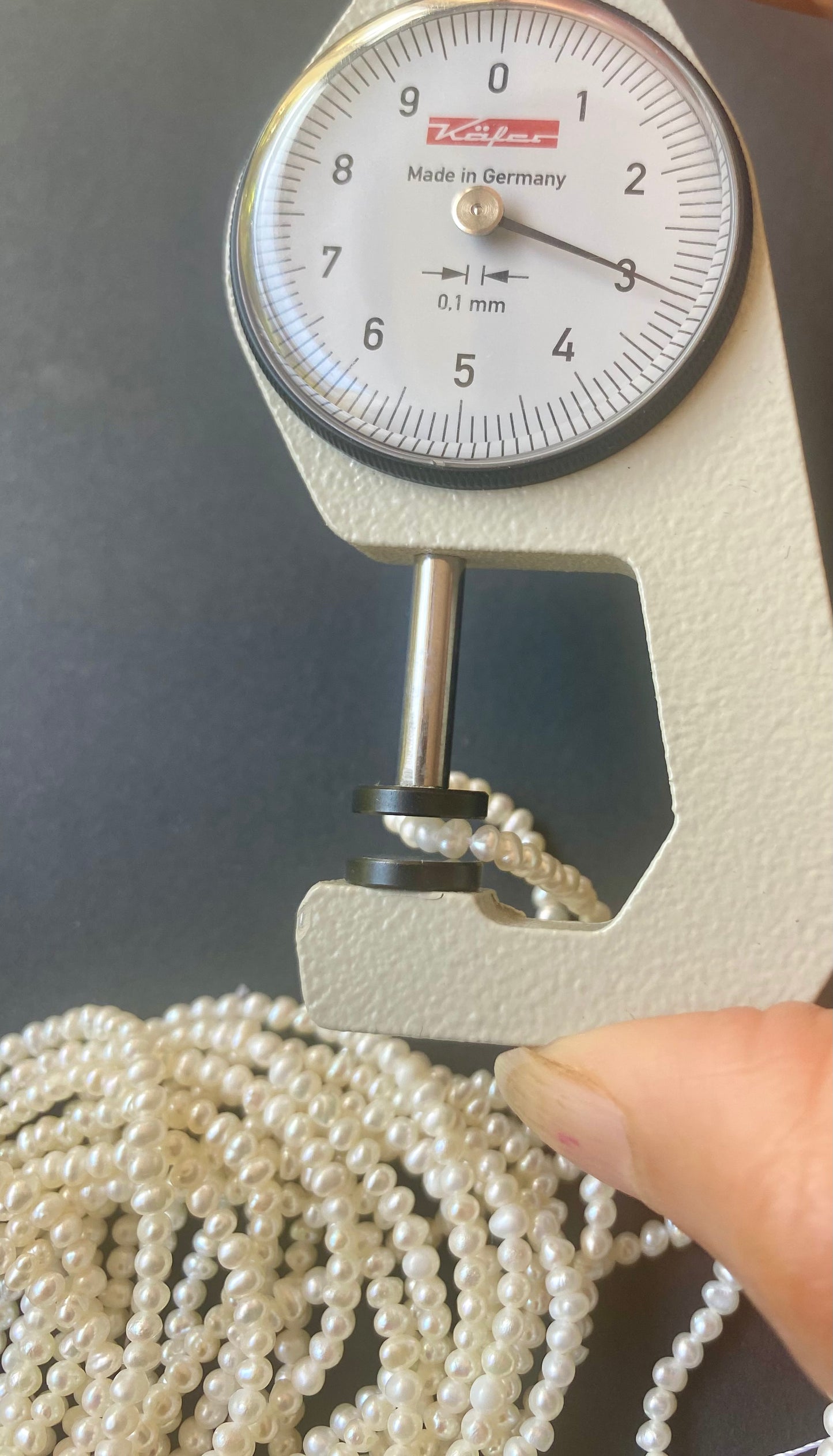 A hand holds a measuring dial gauge next to a pearl necklace crafted from Rondelle Freshwater Pearls.