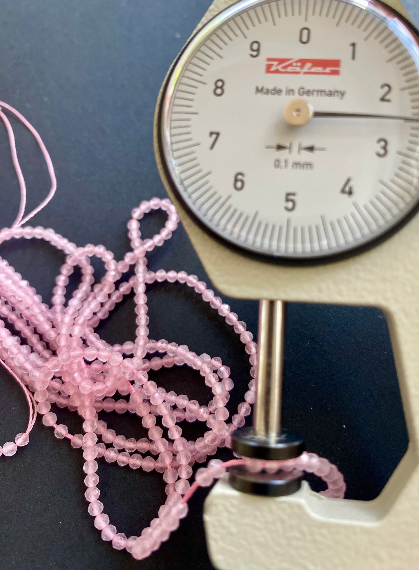 A person measures a pinkish Rose Quartz faceted rondelle stone, 2.5mm, using a dial gauge measuring tool.