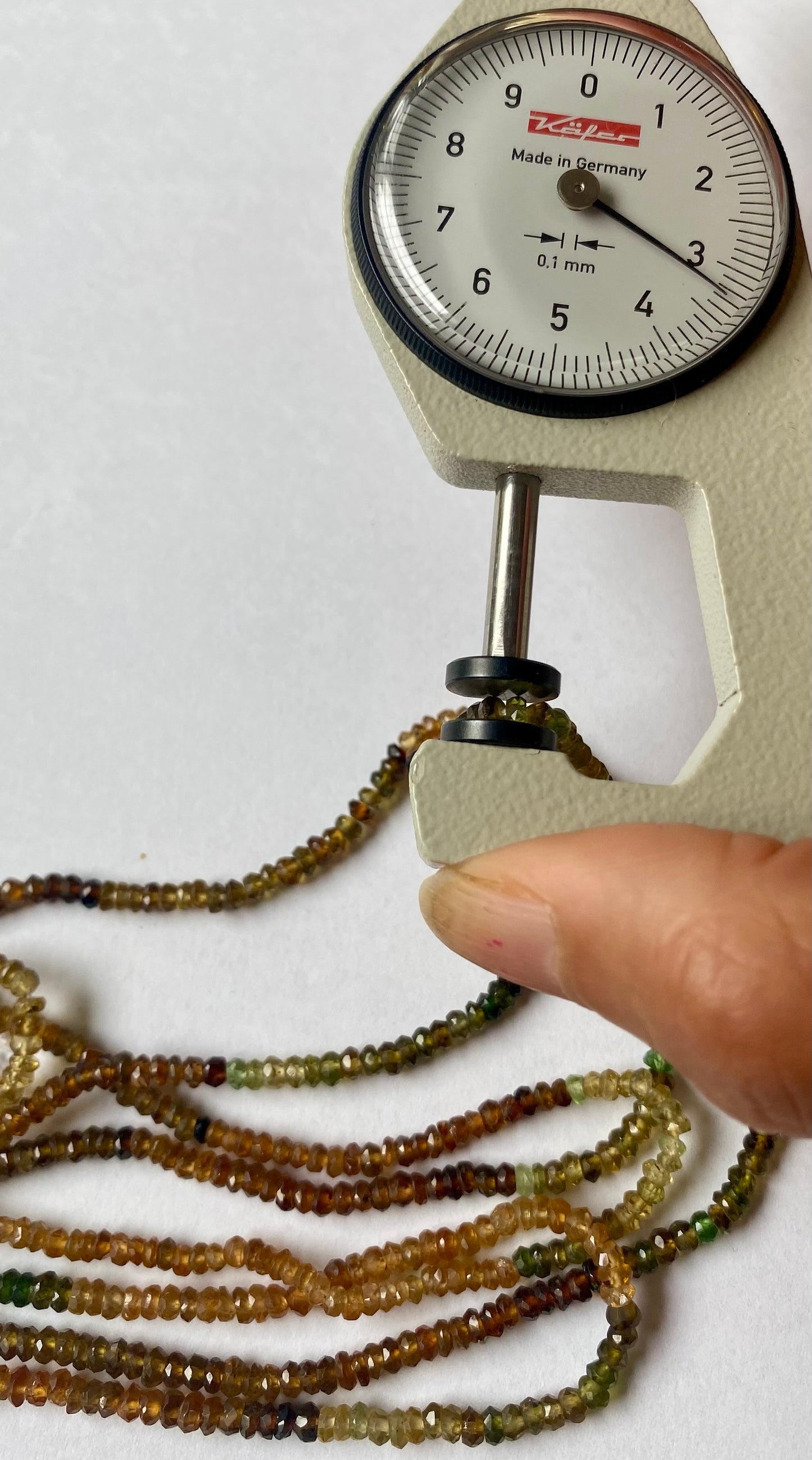A dial gauge next to an array of colourful 3-3.5mm tourmaline beads, showcasing their size and measurement.