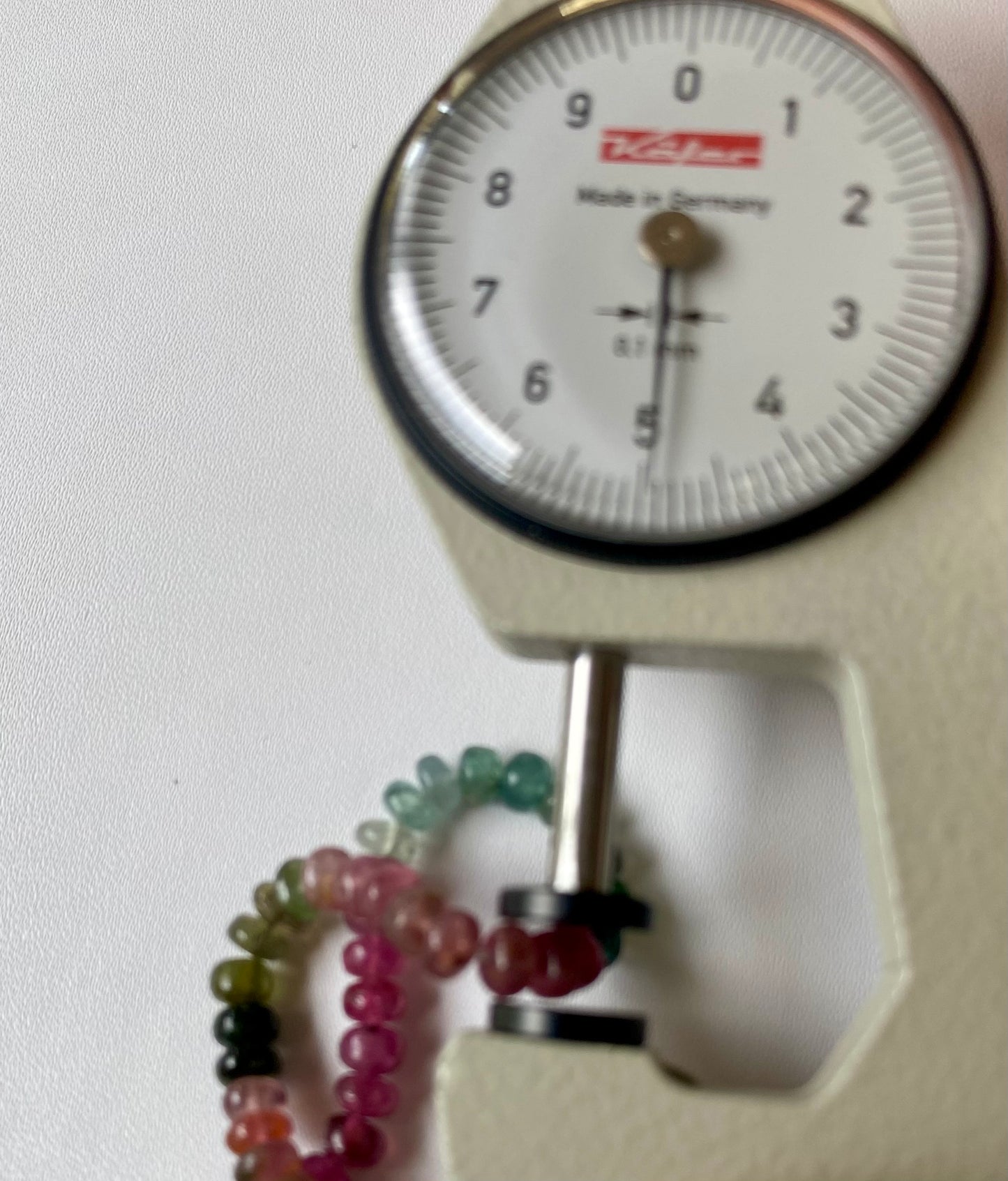 A dial gauge next to an array of colourful 5mm smooth tourmaline beads, showcasing their size and measurement.