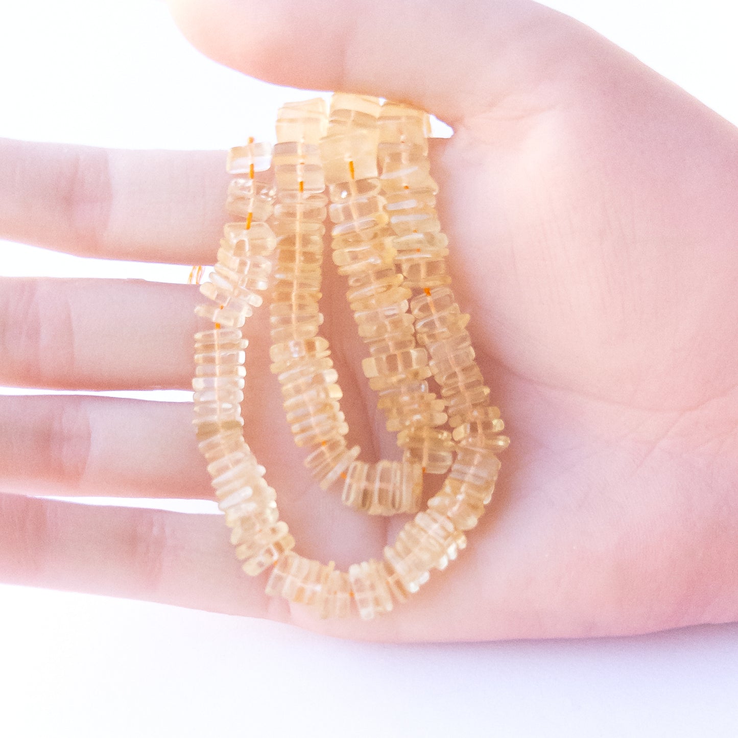 A person holds delicate strands of Citrine beads, showcasing their square heishi design.