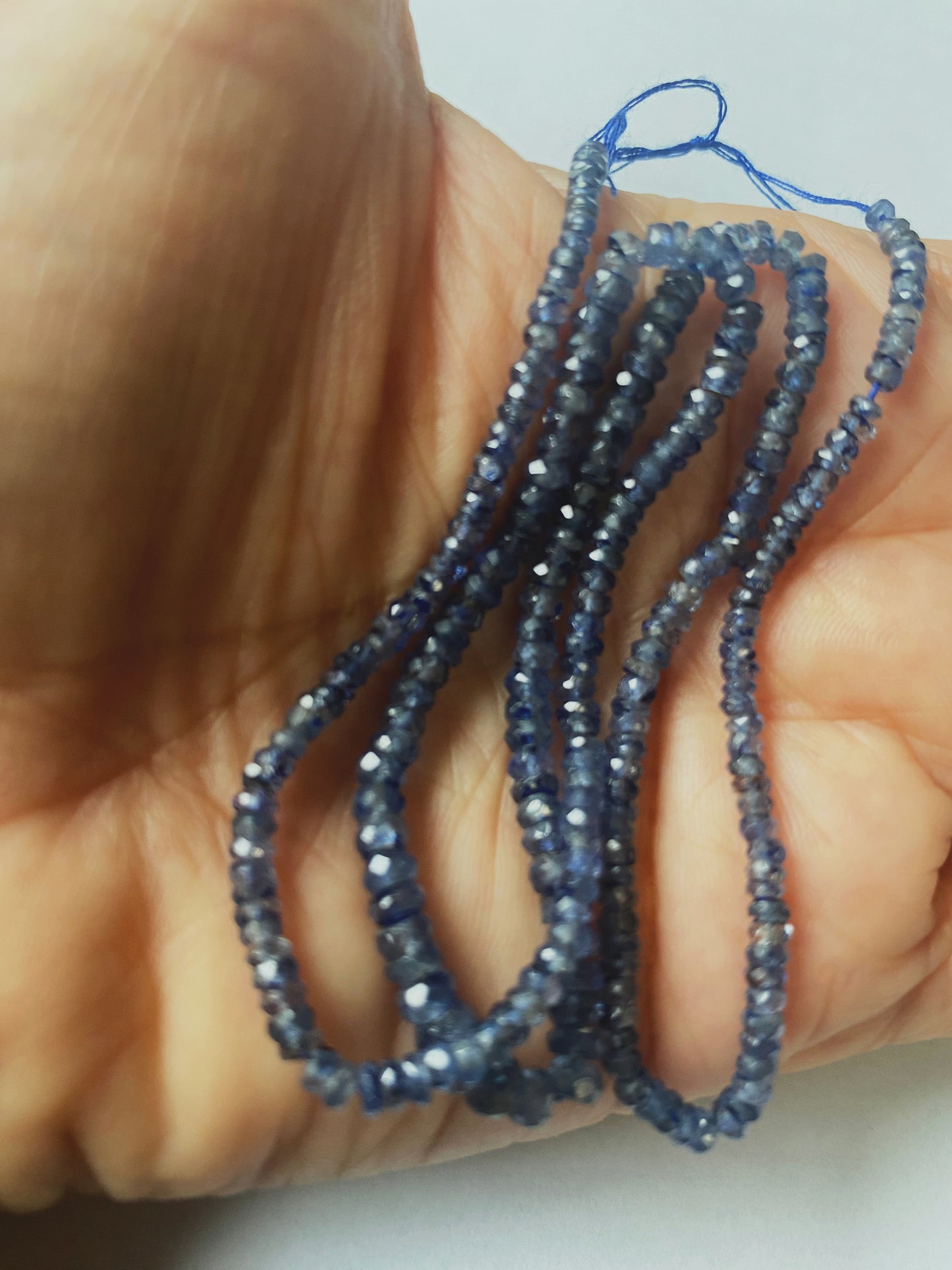 A person holds delicate strands of Denim Blue Sapphire beads, showcasing their smooth design.