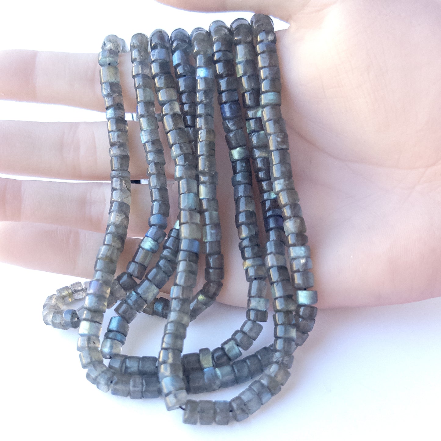 A person holds delicate strands of Labradorite beads, showcasing their smooth cylinder design.