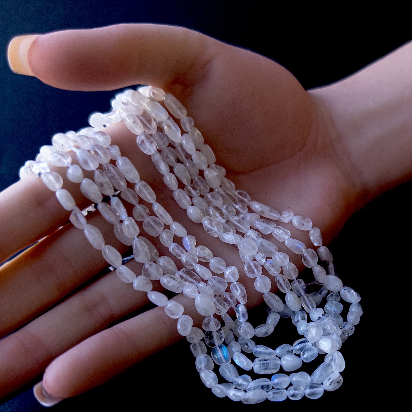 A person holds delicate strands of Moonstone beads, showcasing their smooth tumble design.
