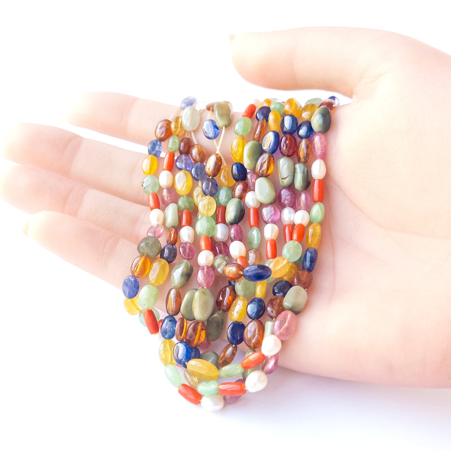 A person holds delicate strands of Multi Precious Navratan beads, showcasing their smooth oval design.
