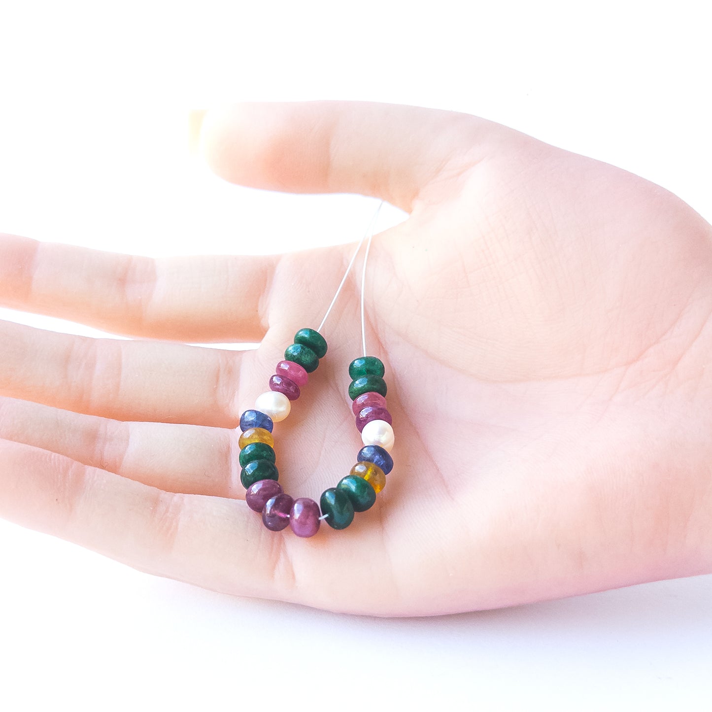 A person holds delicate strands of Ruby, Emerald, Sapphire and Pearl smooth rondelle beads, showcasing their polished design.