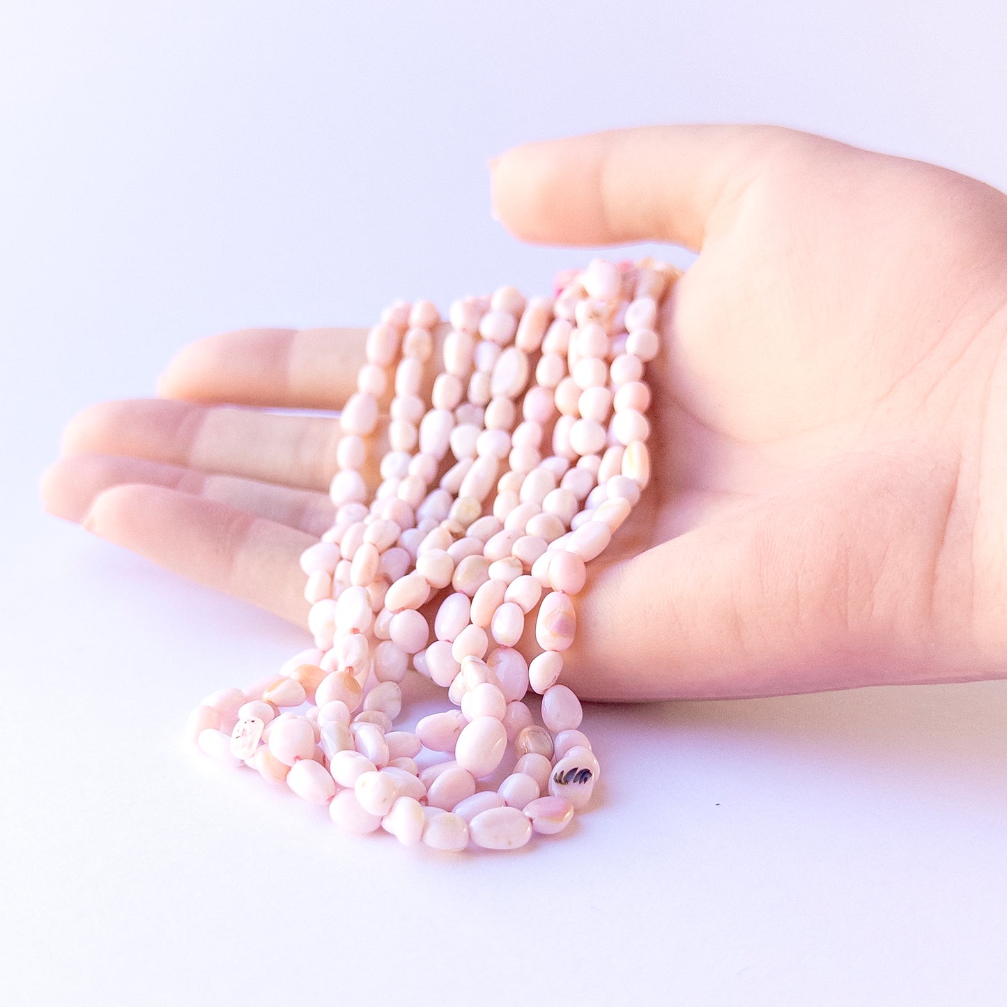 A person holds delicate strands of Pink Opal beads, showcasing their Smooth Tumble design.