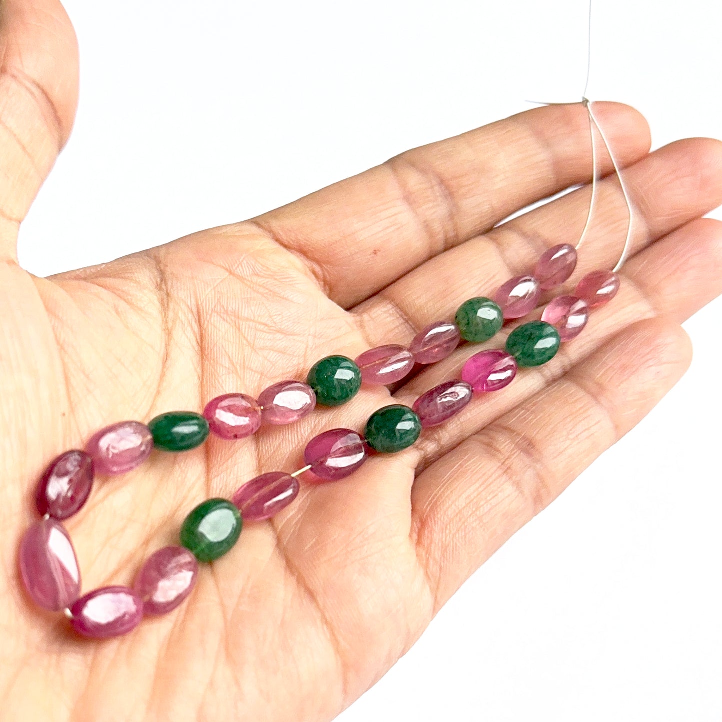 A person holds delicate strands of ruby and emerald oval beads, showcasing their smooth design.