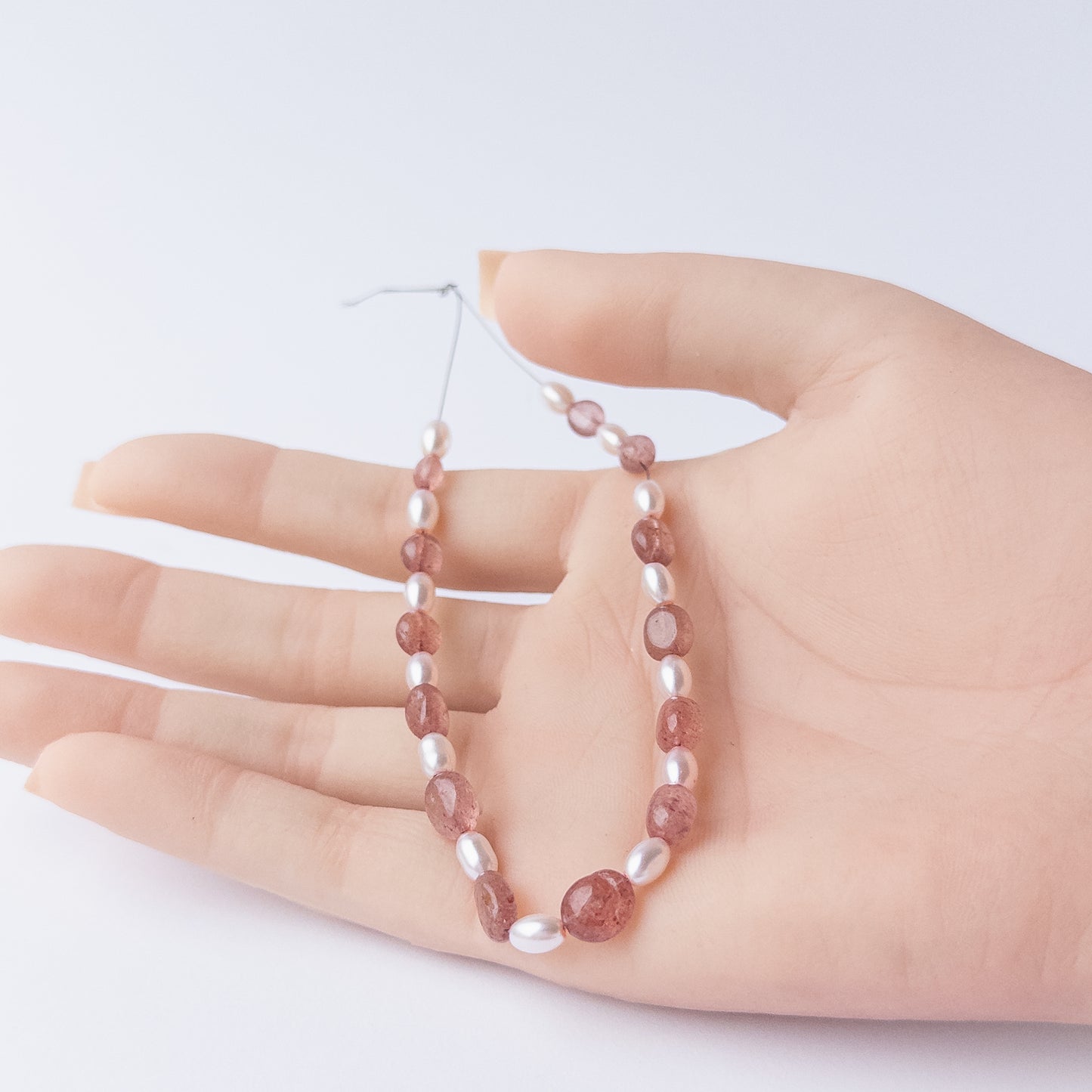 A person holds delicate strands of Strawberry Quartz and Shell Pearl beads, showcasing their smooth oval design.