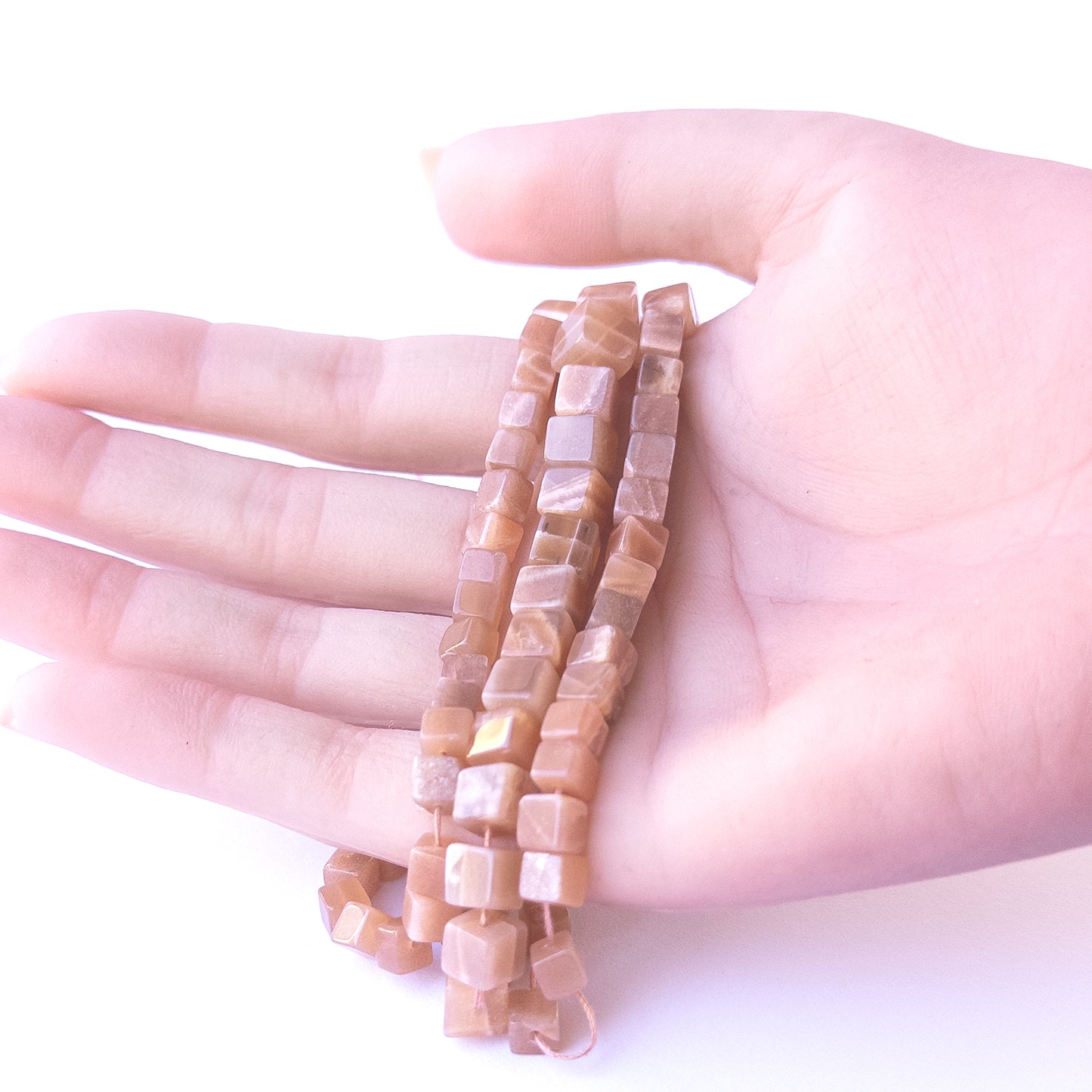 A person holds delicate strands of Sunstone beads, showcasing their smooth cube design.