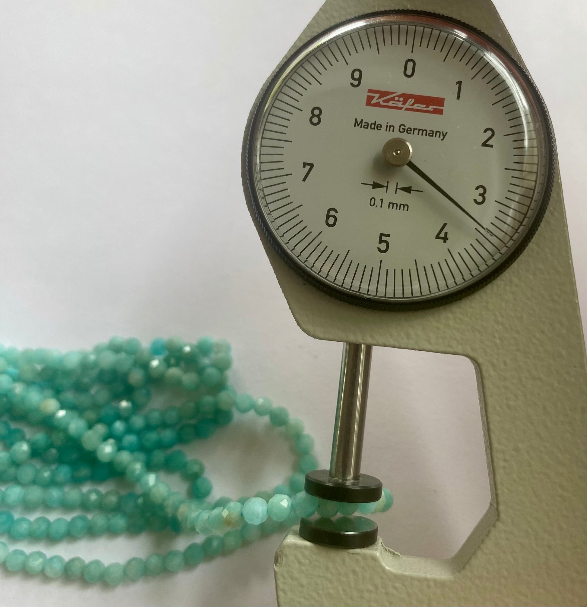 A dial gauge displaying a green blue bead beside a gemstone strand made of faceted rondelle amazonite beads, 3.4-3.5mm in size.