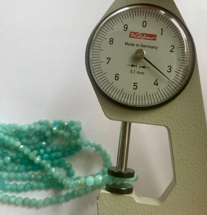 A dial gauge displaying a green blue bead beside a gemstone strand made of faceted rondelle amazonite beads, 3.4-3.5mm in size.