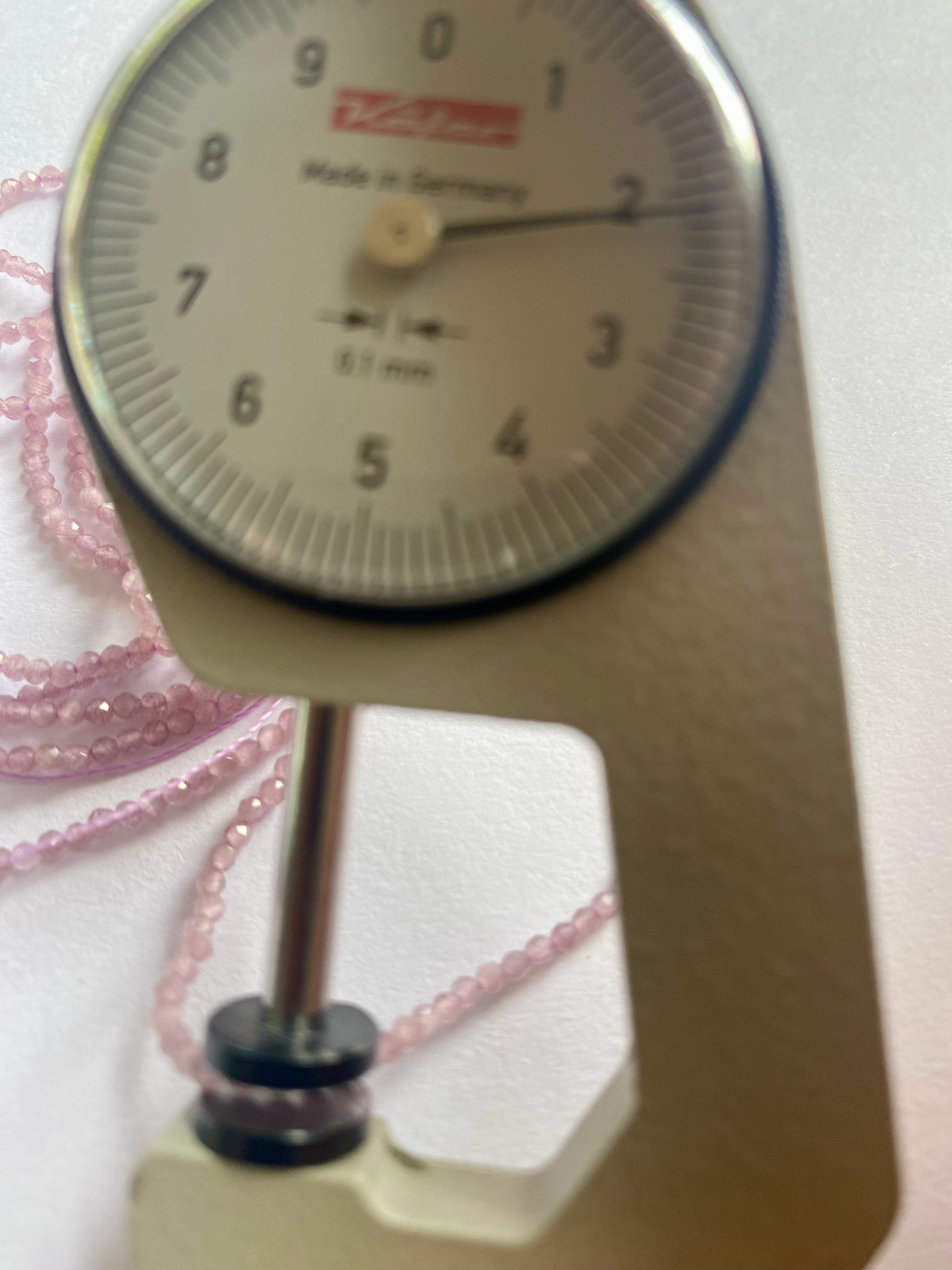 A person measures a purple amethyst rondelle faceted stone, ranging from 2mm to 2.2mm, using a dial gauge measuring tool.