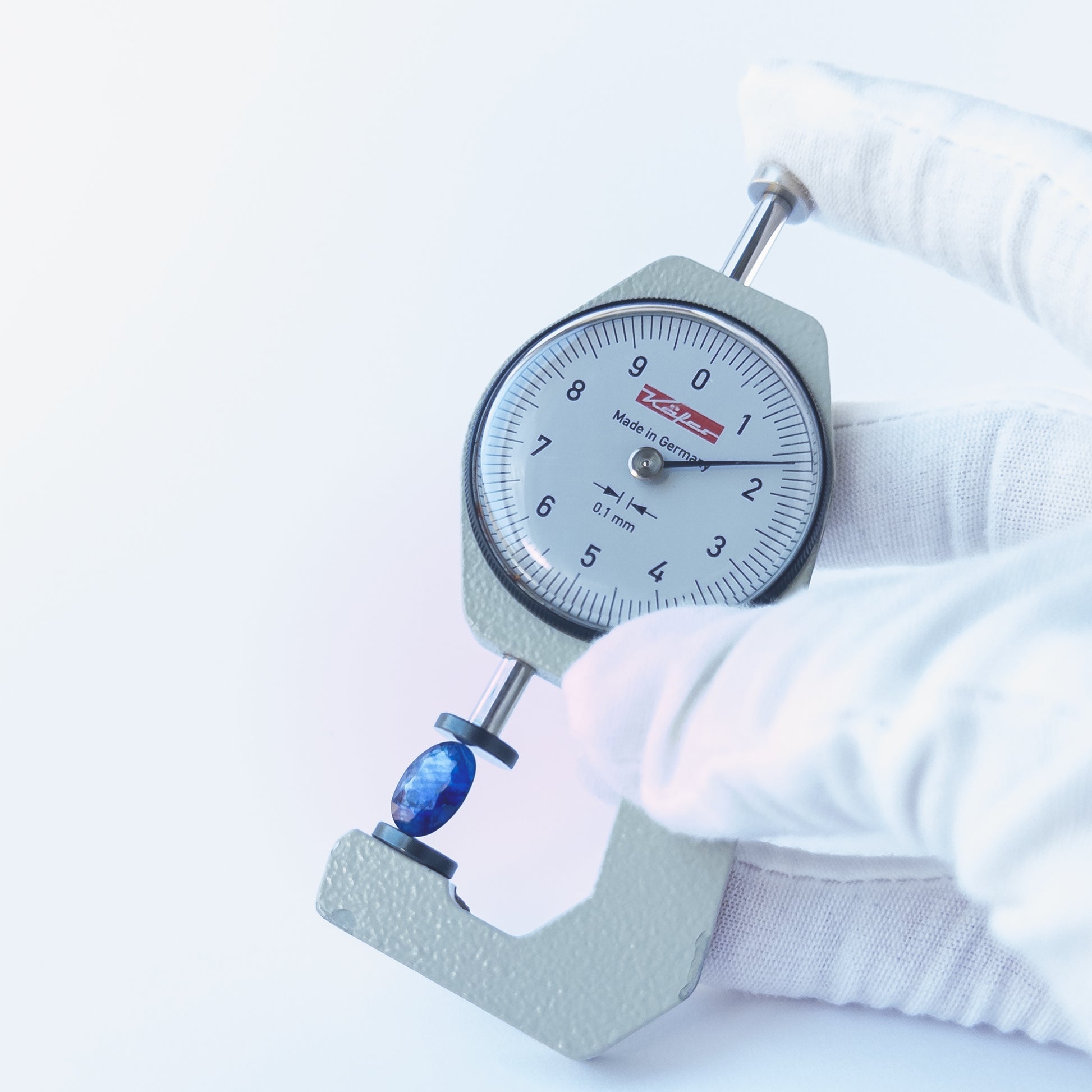 A dial gauge measures the Blue Sapphire faceted oval 11.5mm × 8.4mm gemstone.