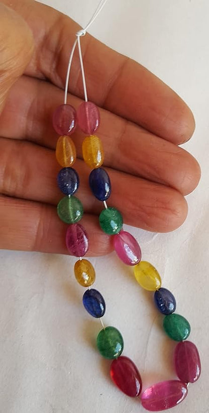 A person holds a vibrant bunch of multi-coloured smooth oval beads, including ruby, blue sapphire, yellow sapphire, and emerald.
